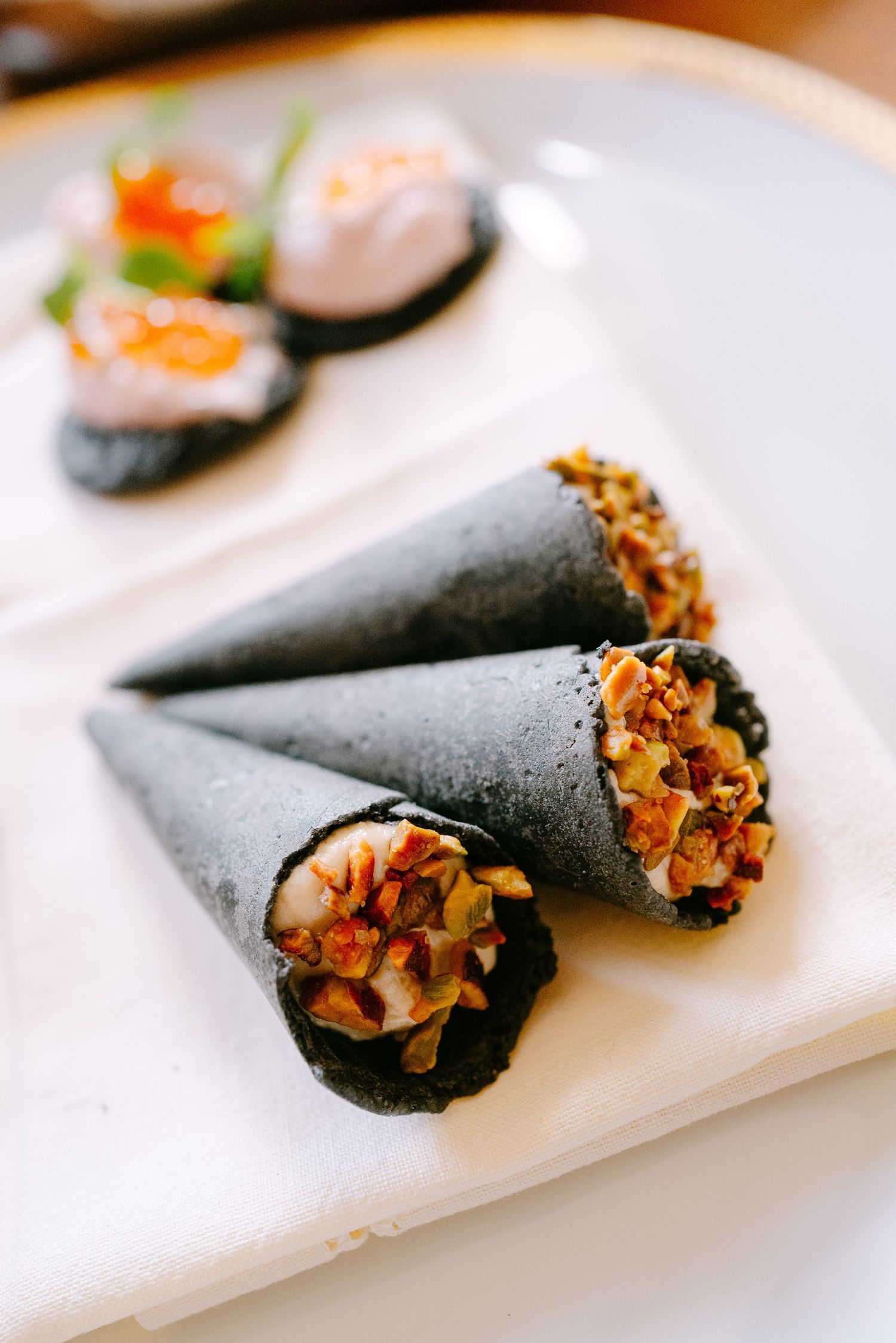Black squid ink cones filled with savory toppings served on a white plate with sushi bites in background.