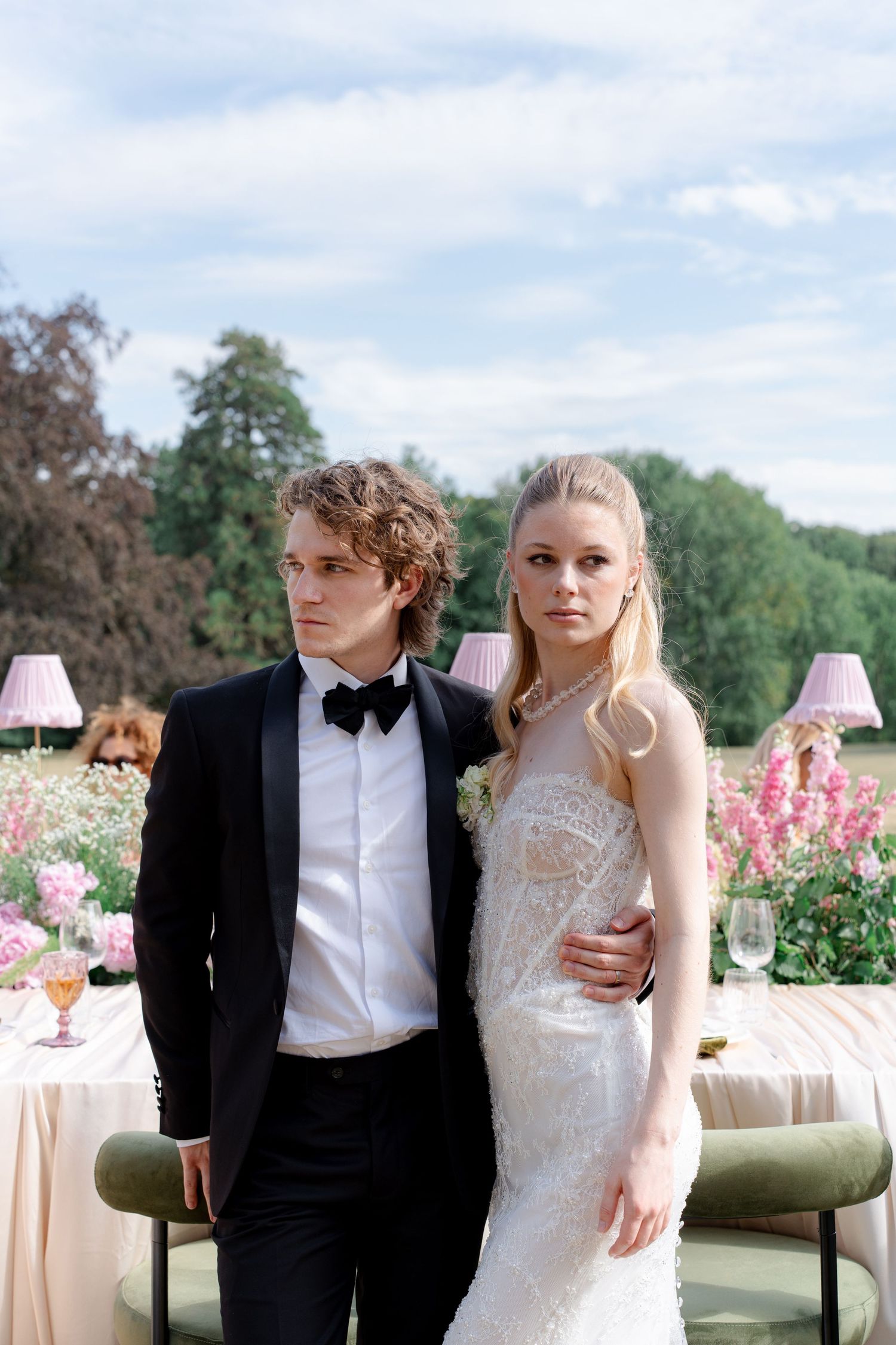 Elegant bride and groom posing outdoors at a floral wedding reception, wearing white lace gown and black tuxedo.