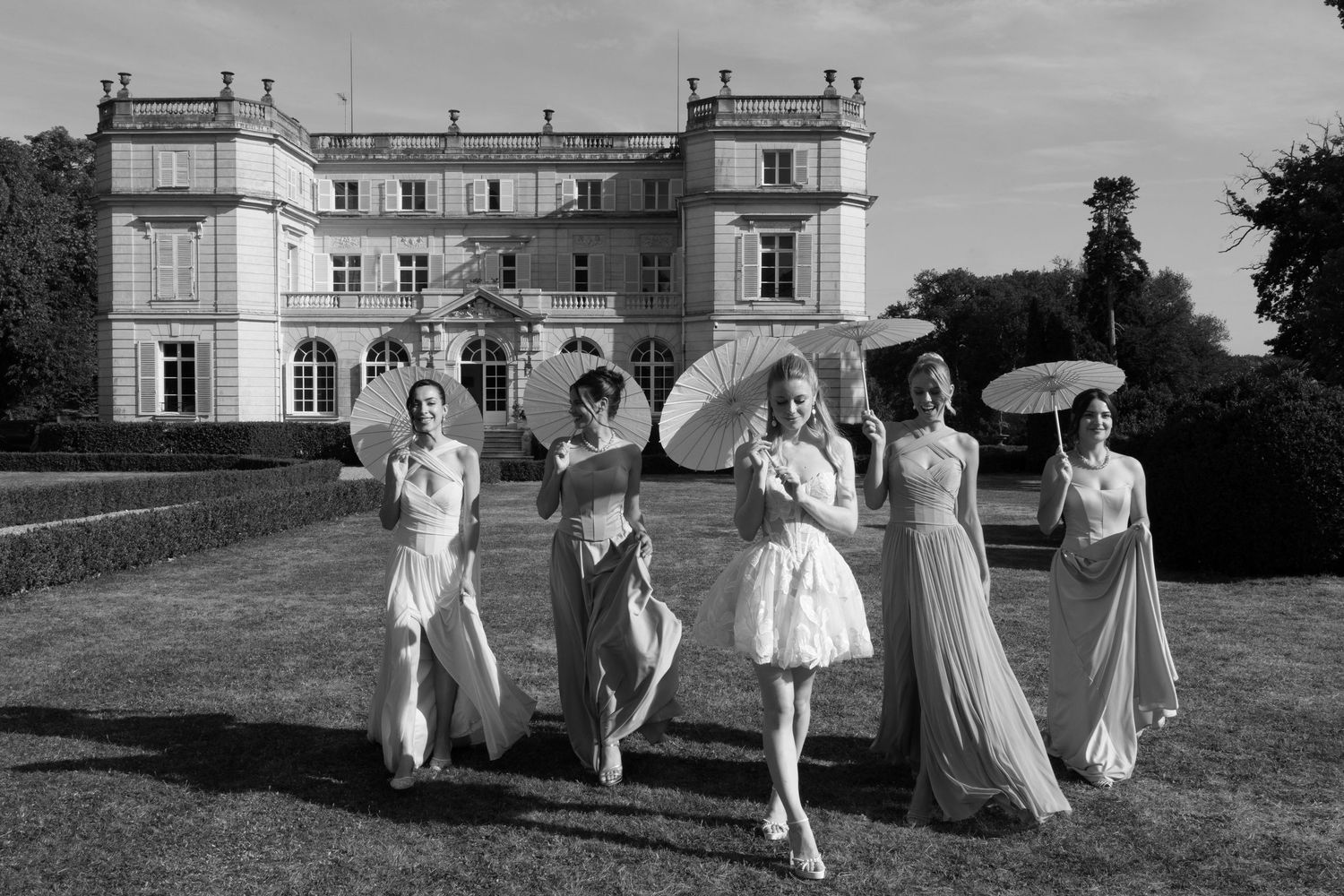Black and white photo of five elegant women in gowns holding parasols walking in front of a grand French chateau.