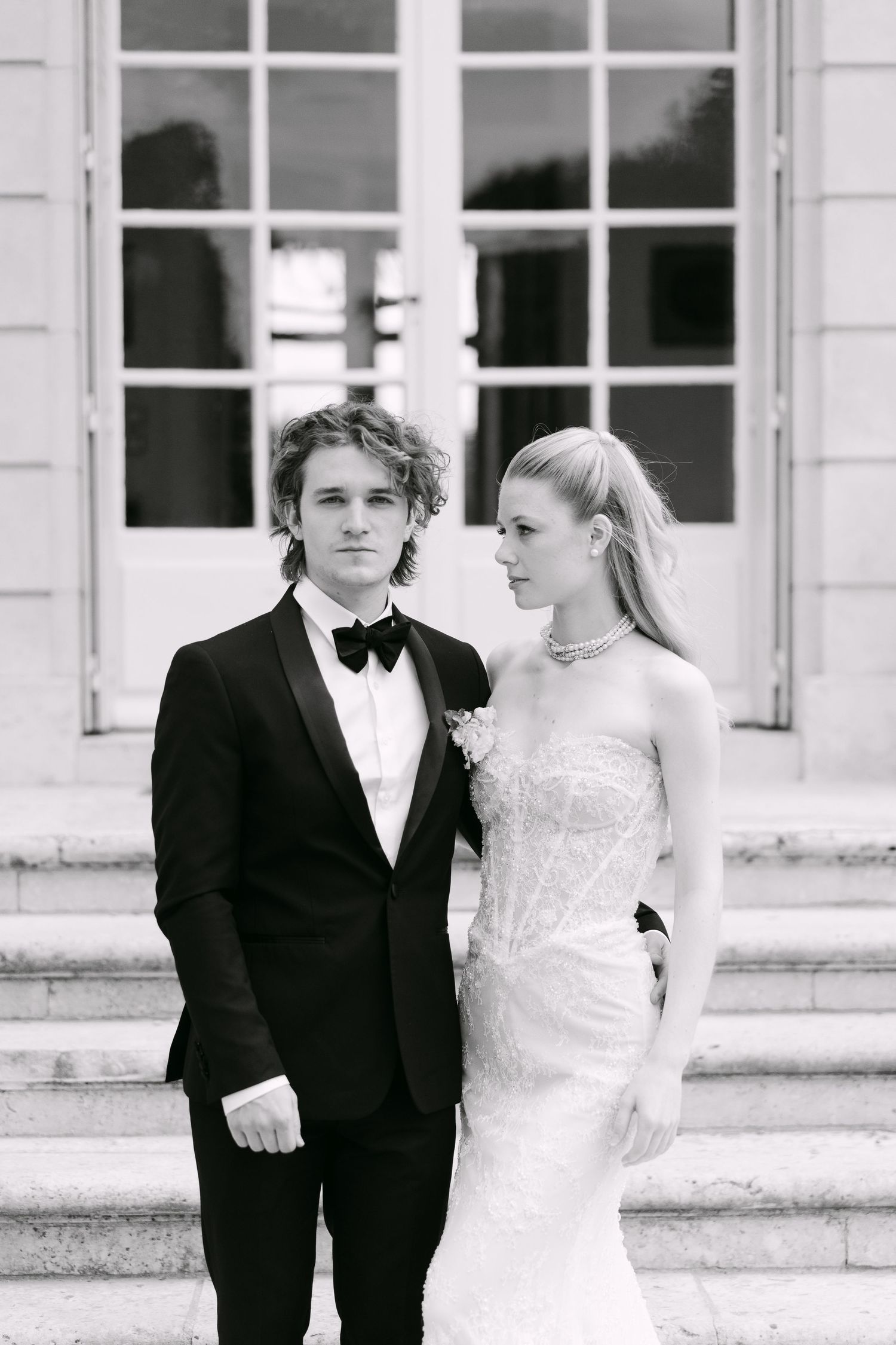 Black and white wedding portrait of bride in strapless lace gown and groom in tuxedo on elegant stone steps.