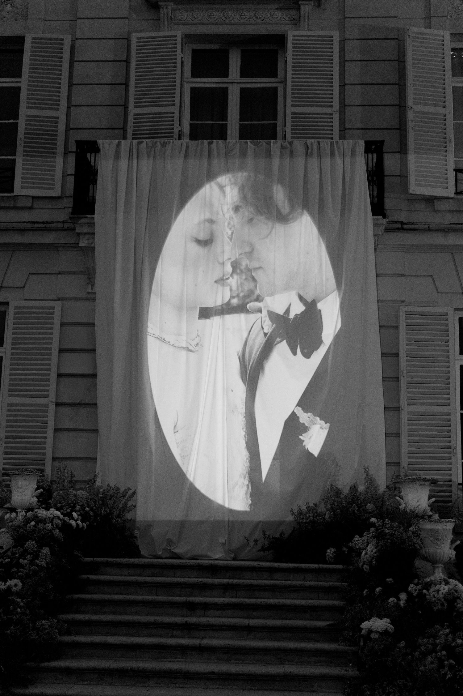 Black and white photo of a romantic couple kissing, projected onto a draped curtain on a classic French building facade.
