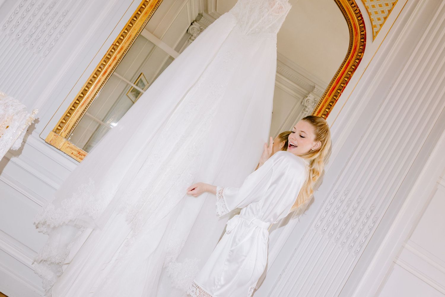 Bride in white robe admiring her wedding dress hanging near an ornate gold mirror in an elegant room.