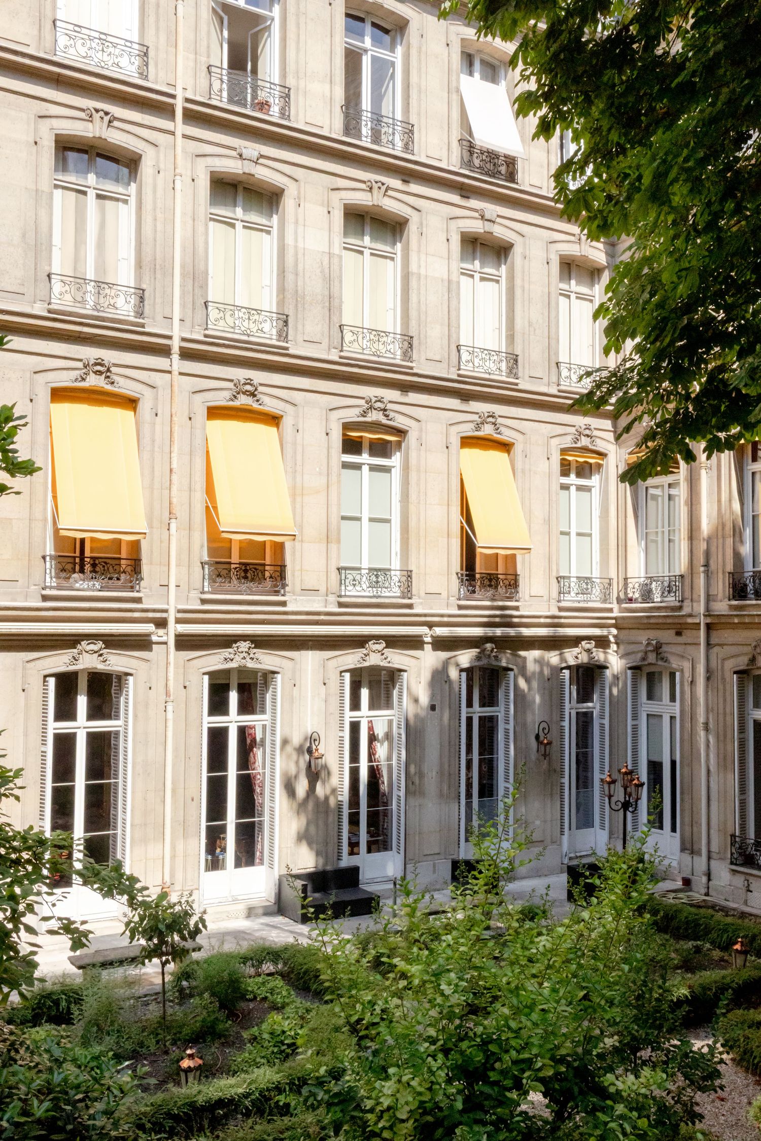 Elegant Haussmann-style Parisian building with yellow awnings, ornate balconies, and lush garden courtyard.
