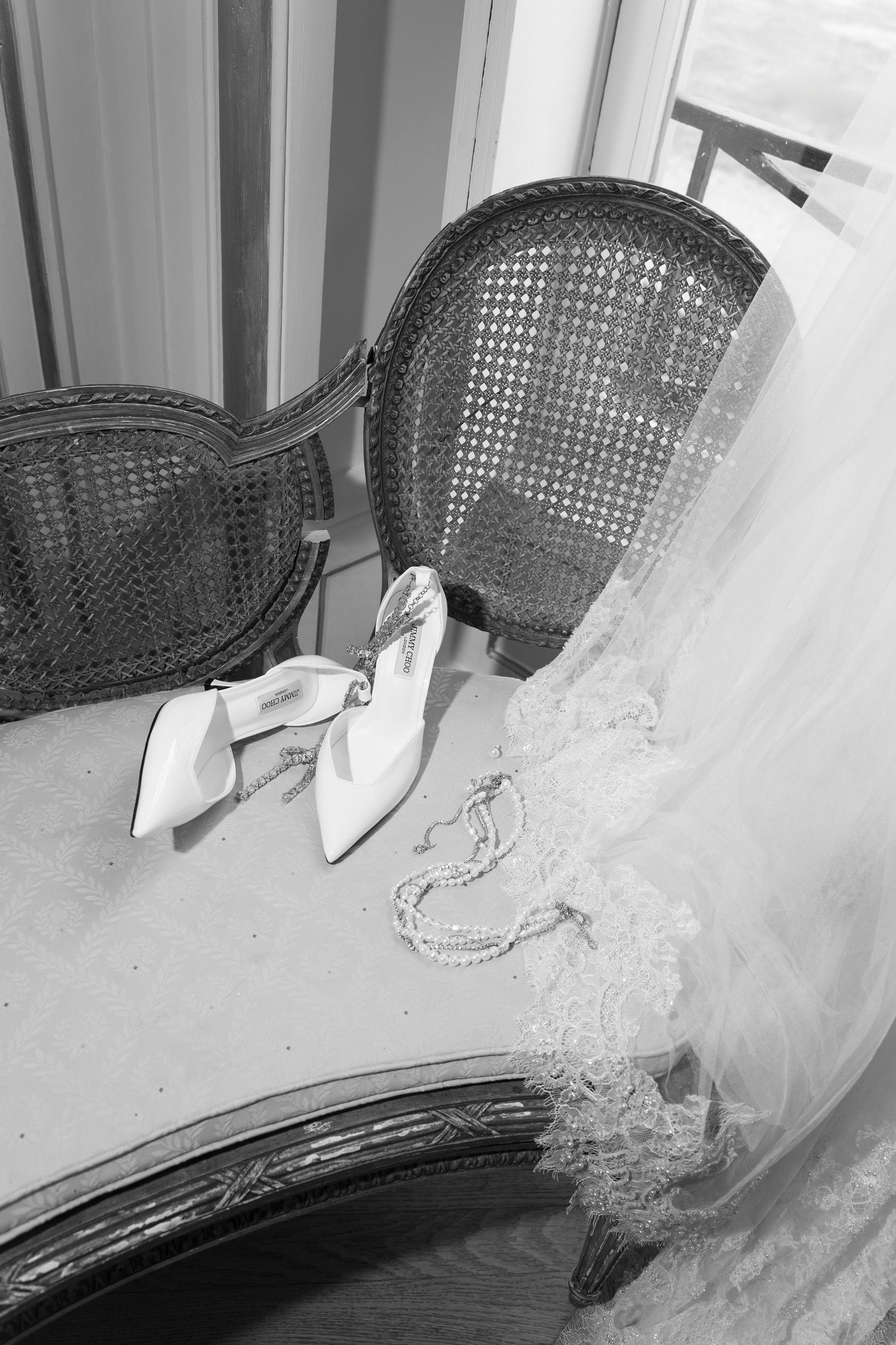 Black and white photo of bridal heels, pearl necklace, and lace veil arranged on vintage table near window.