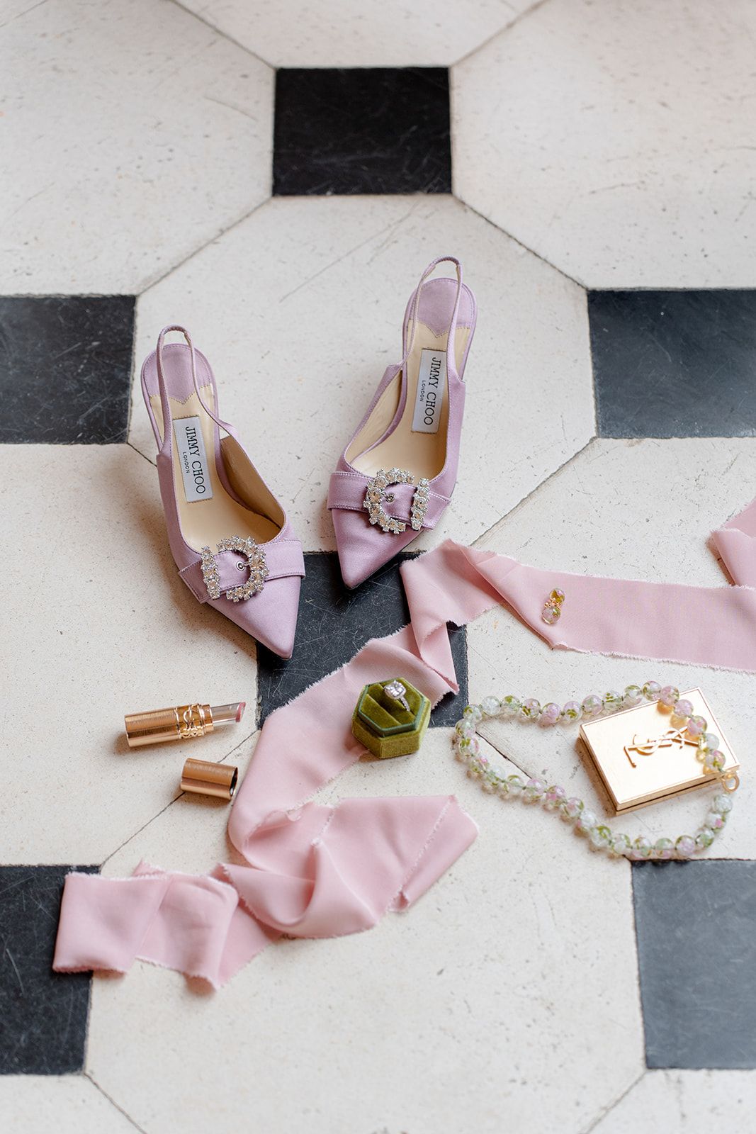 Blush pink Jimmy Choo bridal heels with crystal bow detail styled with ribbon, bracelet, and gold compact on tile.