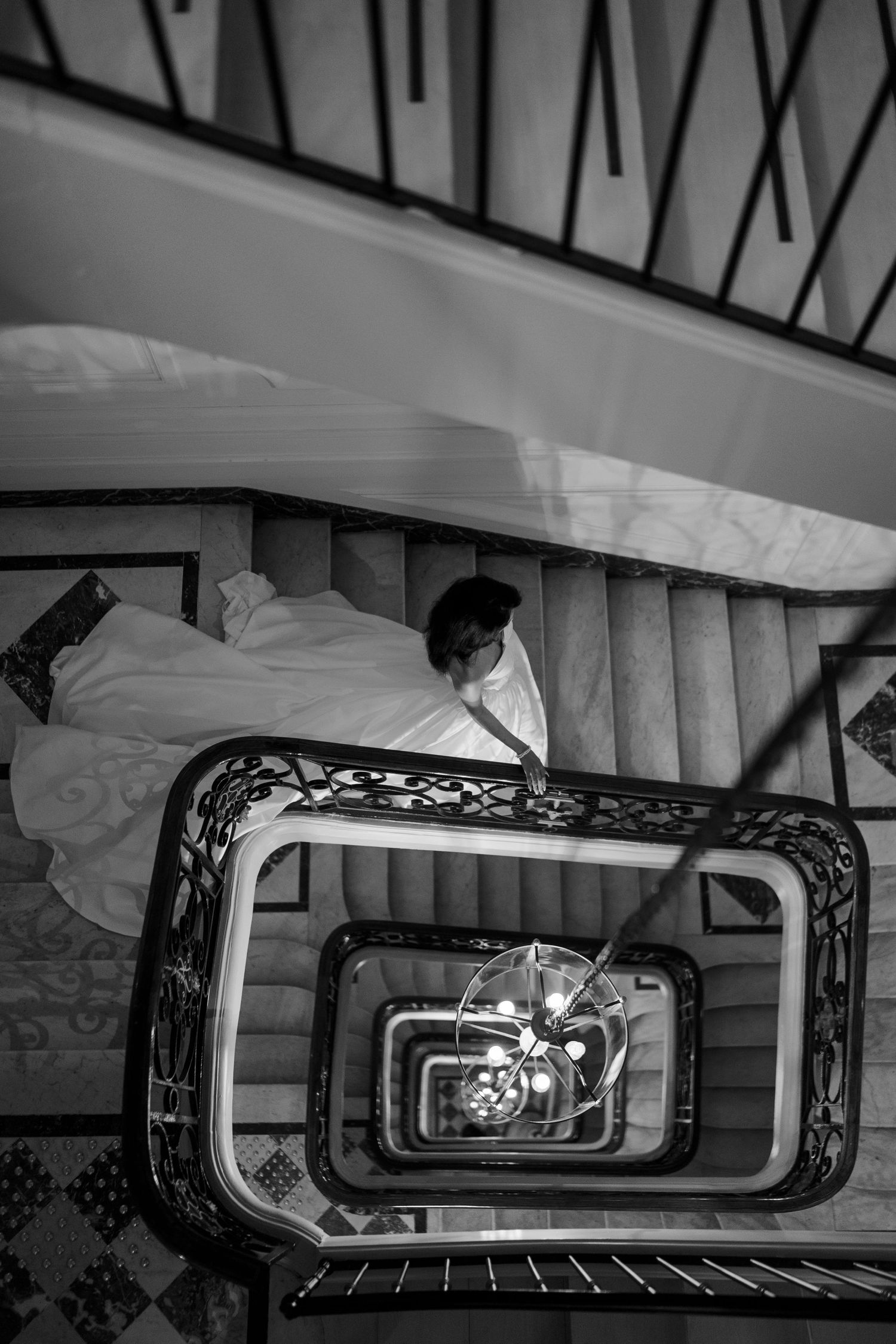 Black and white aerial view of a bride in a flowing gown descending an ornate spiral staircase.