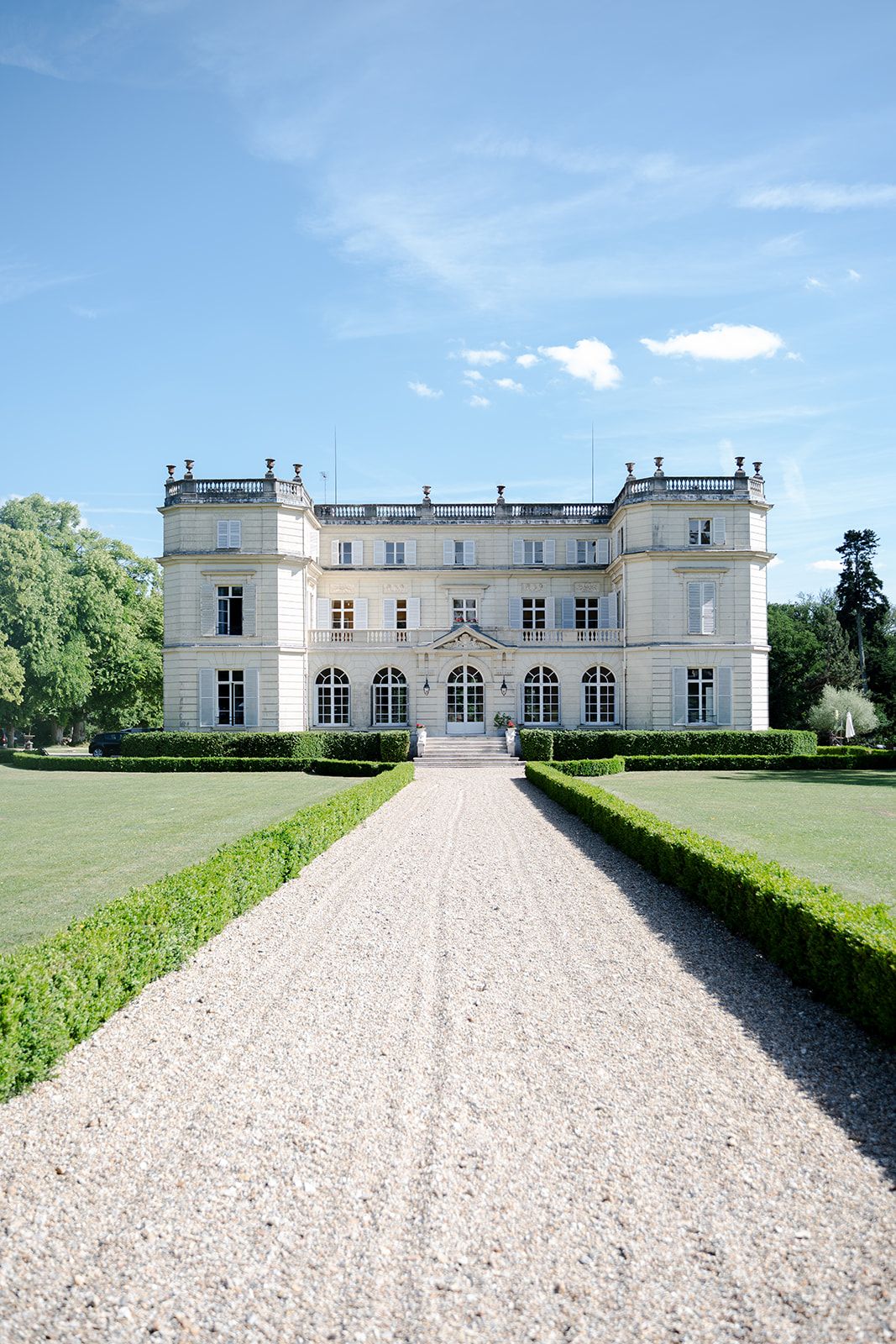 Grand French chateau with gravel driveway, symmetrical boxwood hedges, and arched windows under a sunny sky.