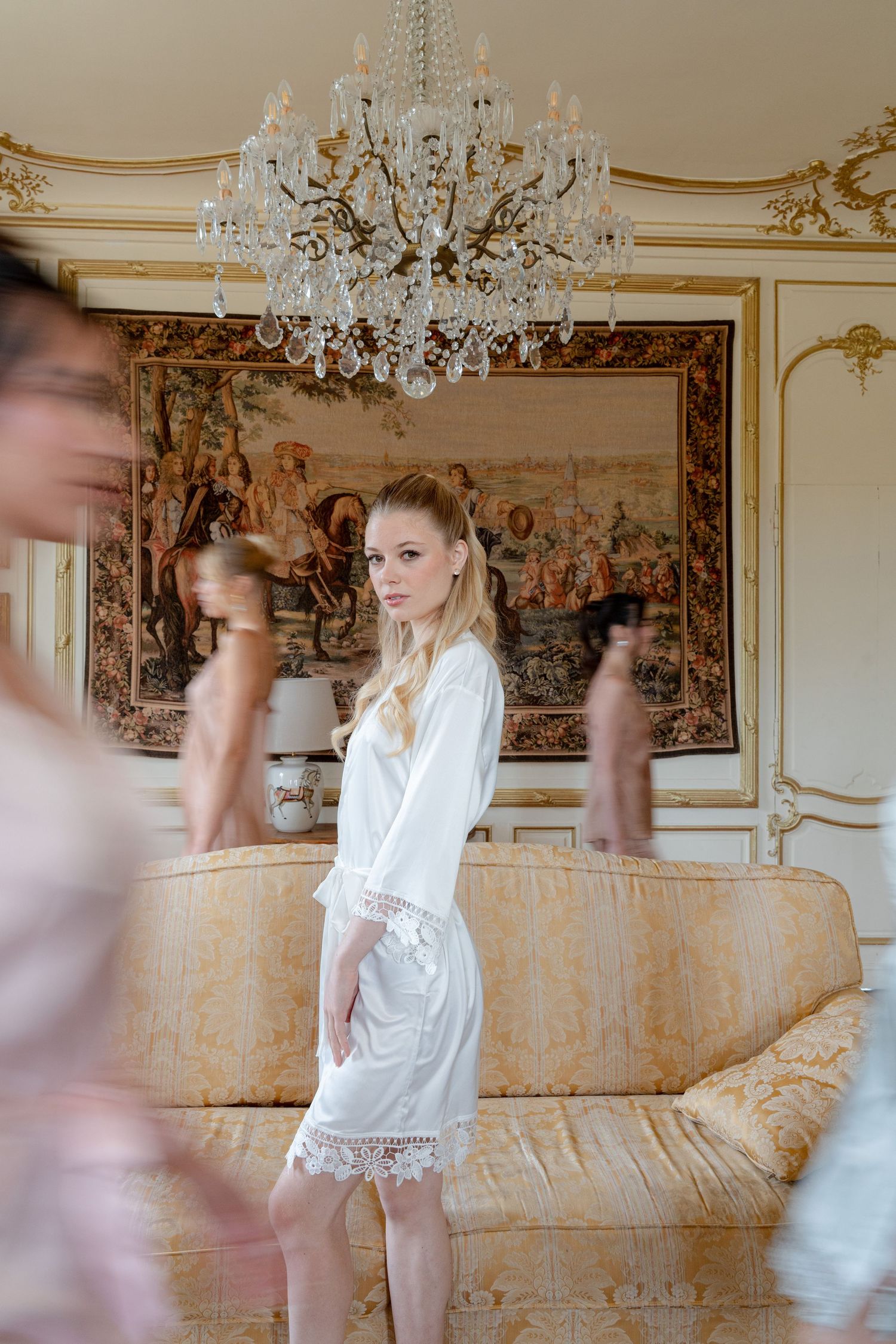 Blonde bride in white lace robe stands in ornate room with crystal chandelier and tapestry.