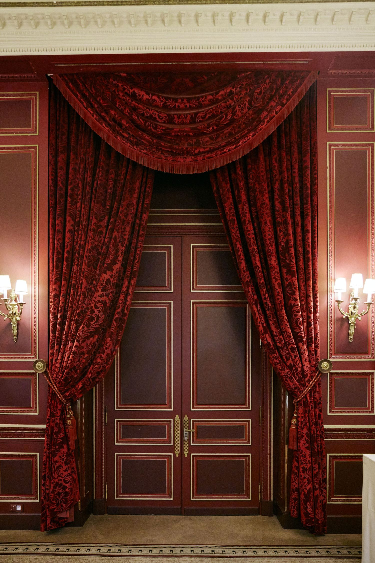 Elegant red damask curtains draped over ornate dark wood double doors with gold wall sconces in a luxurious interior.