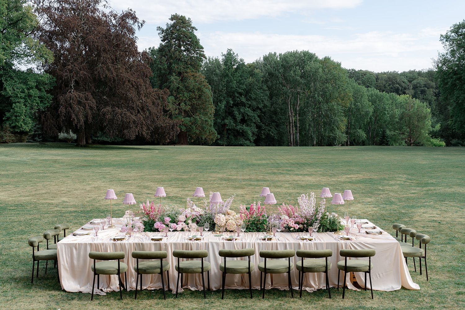 Romantic al fresco wedding dining setup with pink blooms and candles surrounded by tall trees in open field.