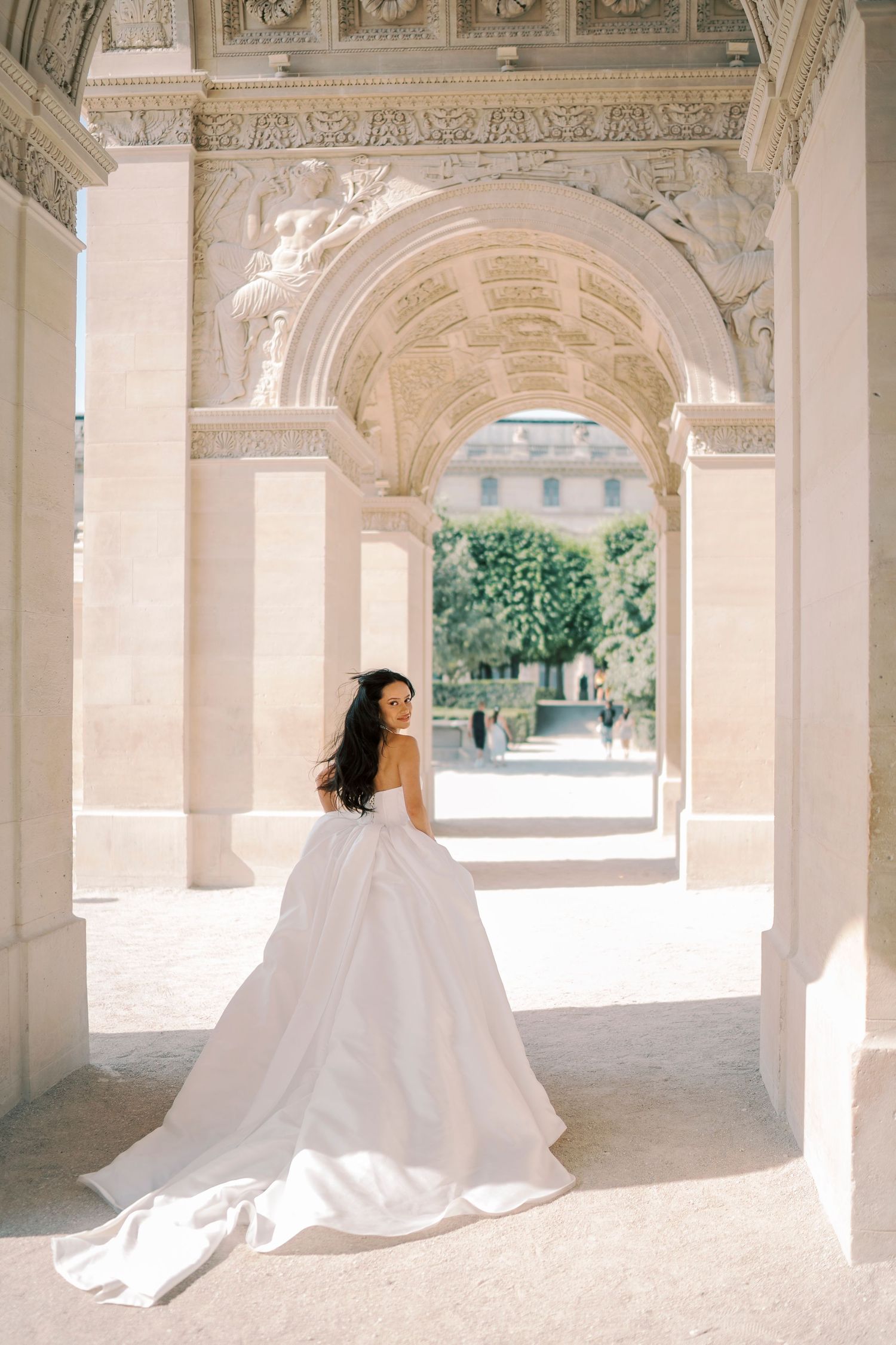 Bride in elegant white ball gown with train poses under ornate stone arches in Paris.