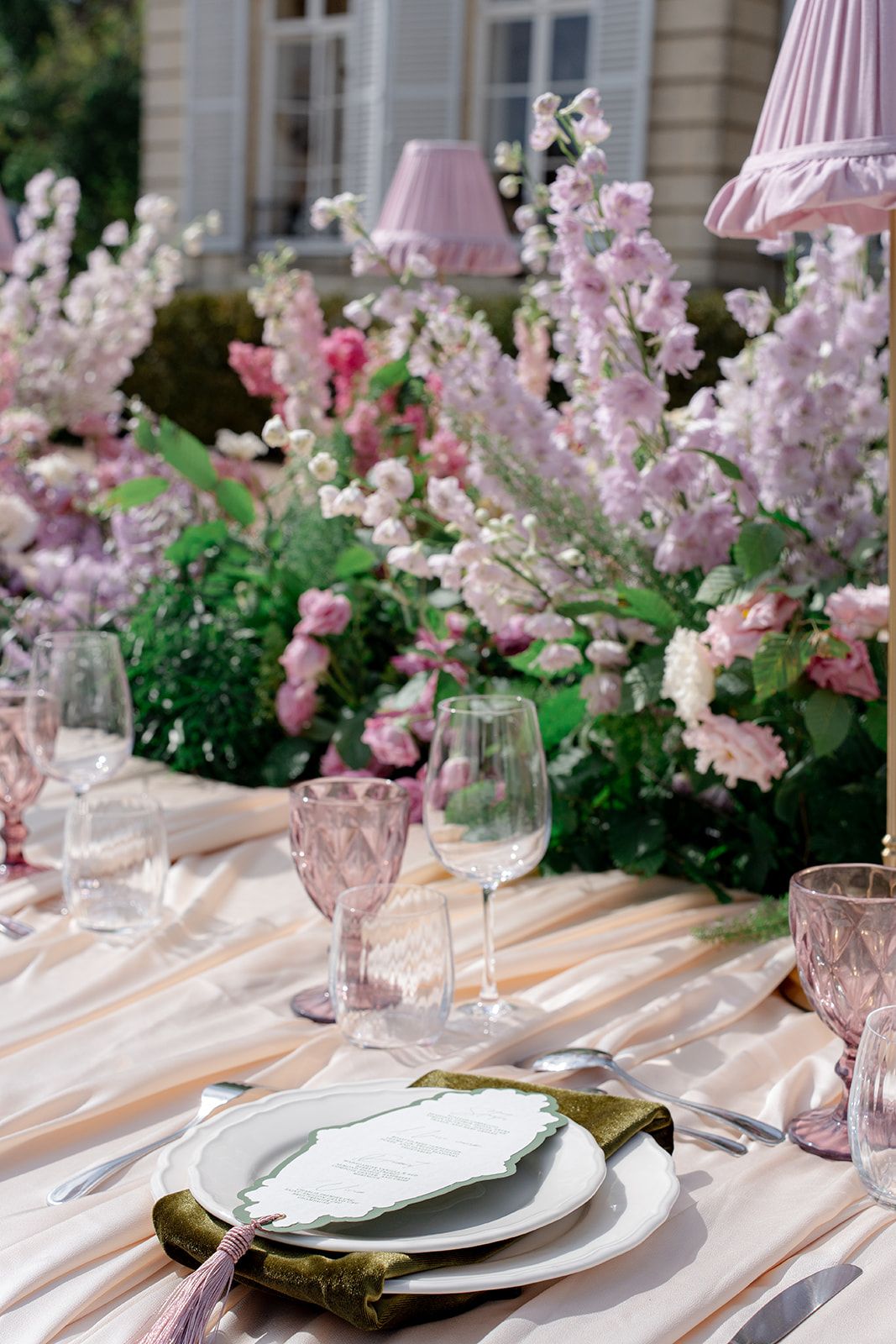 Elegant outdoor wedding place setting with pink glassware, green napkin, and lush pink floral centerpieces.