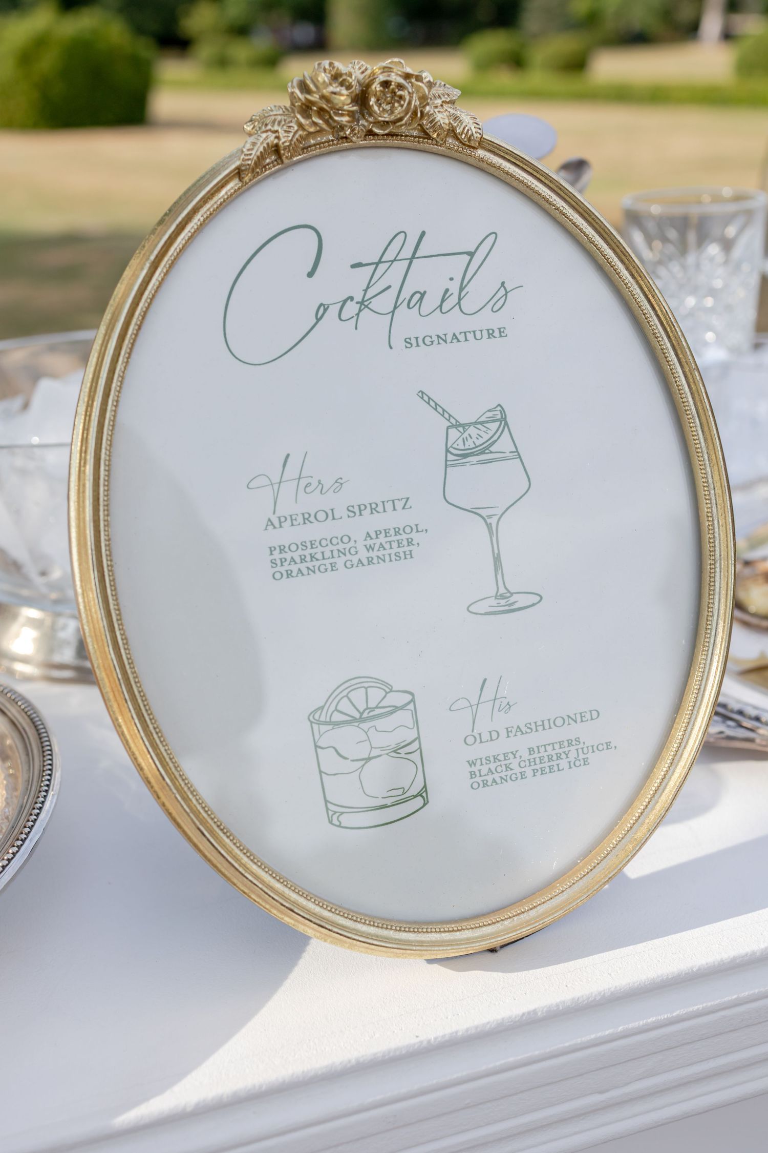 Gold oval framed signature cocktails menu displaying His and Hers drinks at an elegant outdoor wedding reception.