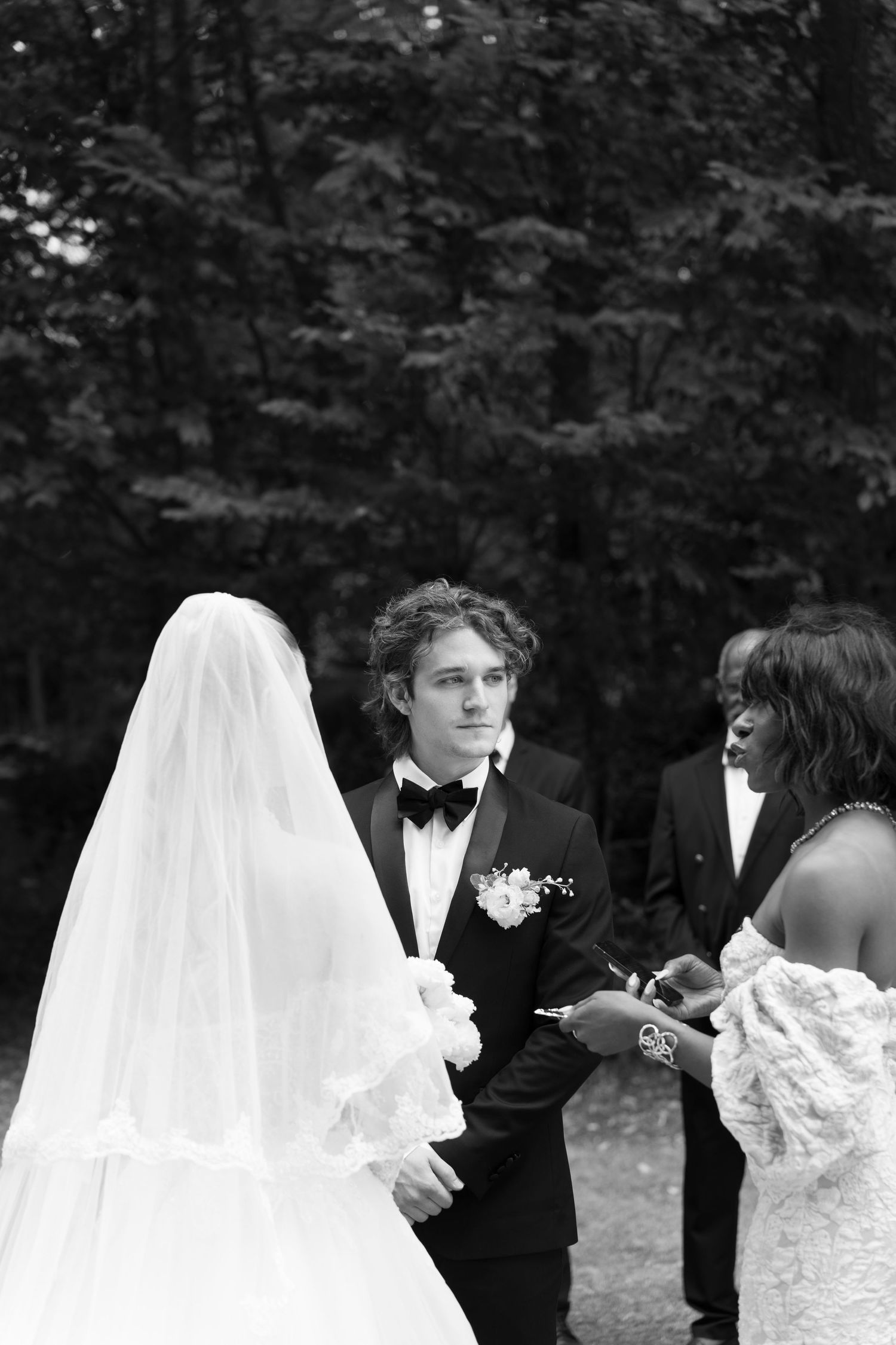 Black and white outdoor wedding ceremony with groom in tuxedo exchanging rings with bride in veil and lace dress.