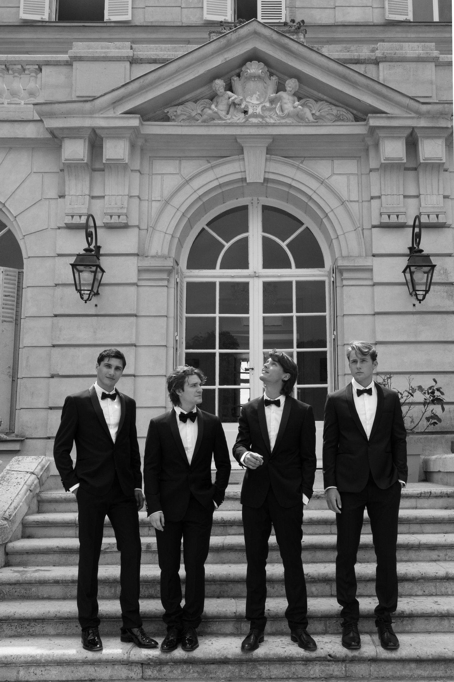 Four young men in black tuxedos and bow ties pose on steps before a grand classical building.