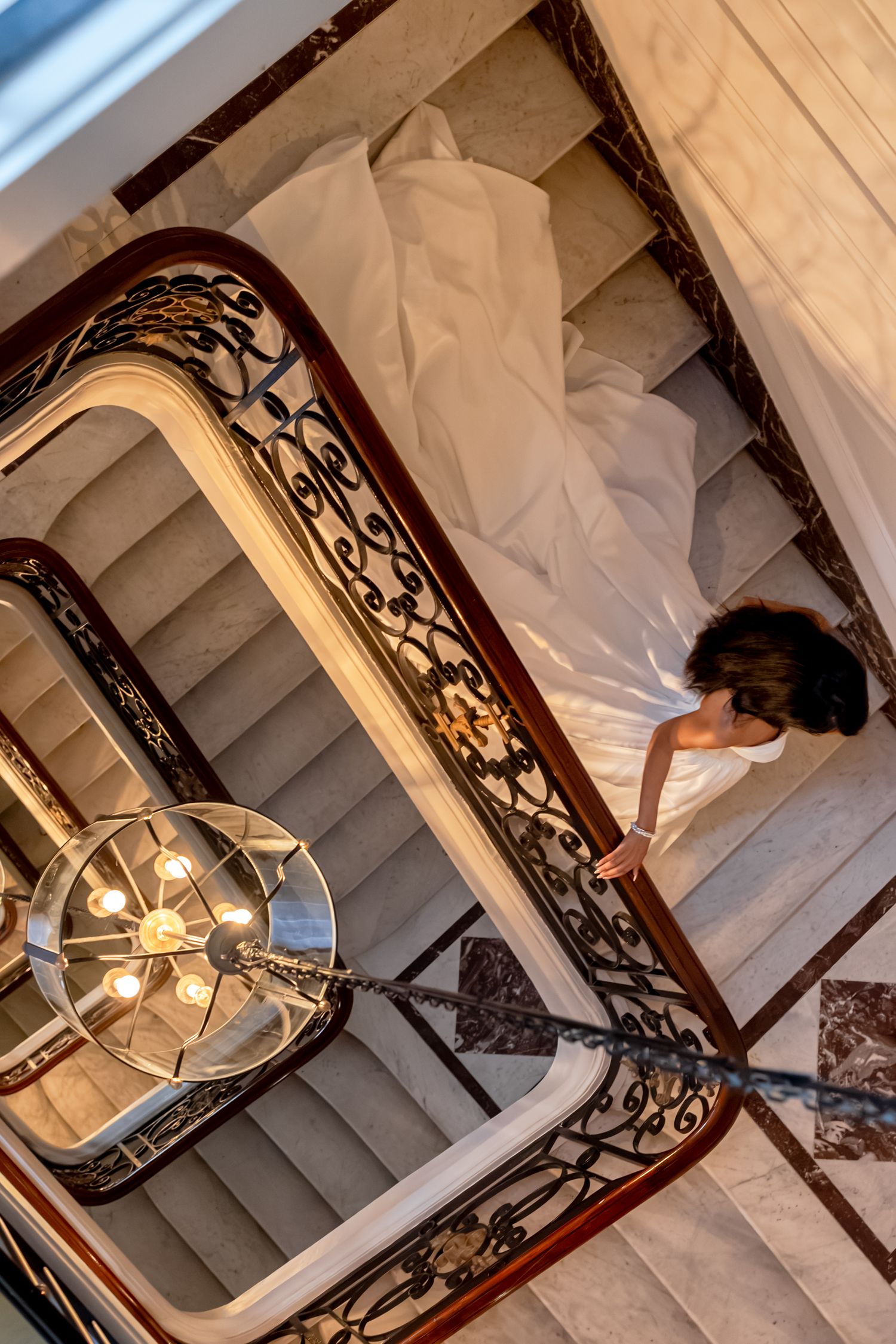 Aerial view of bride in white gown descending ornate marble staircase with wrought iron railings.