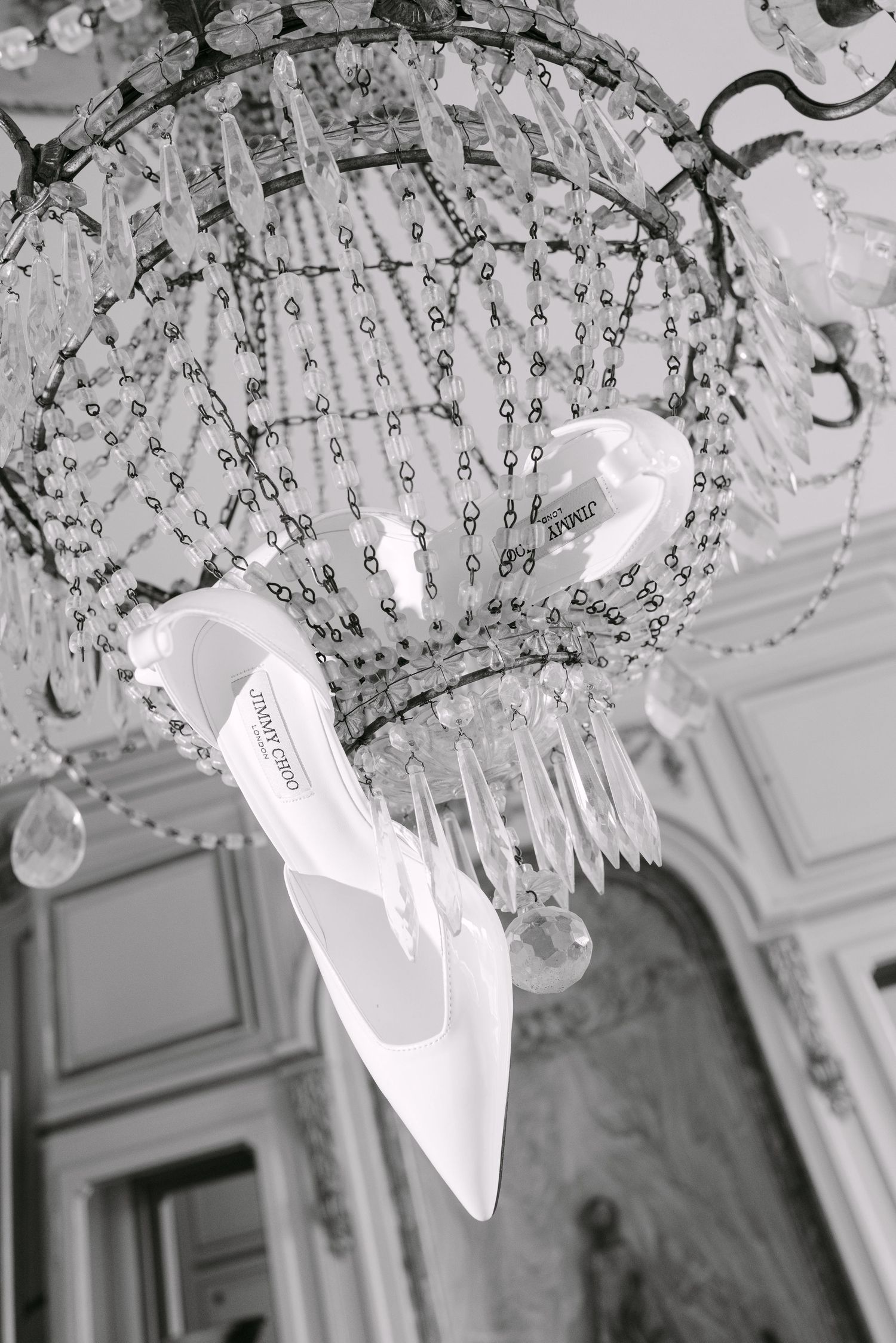 Black and white photo of a white Jimmy Choo bridal heel hanging elegantly from a crystal chandelier.