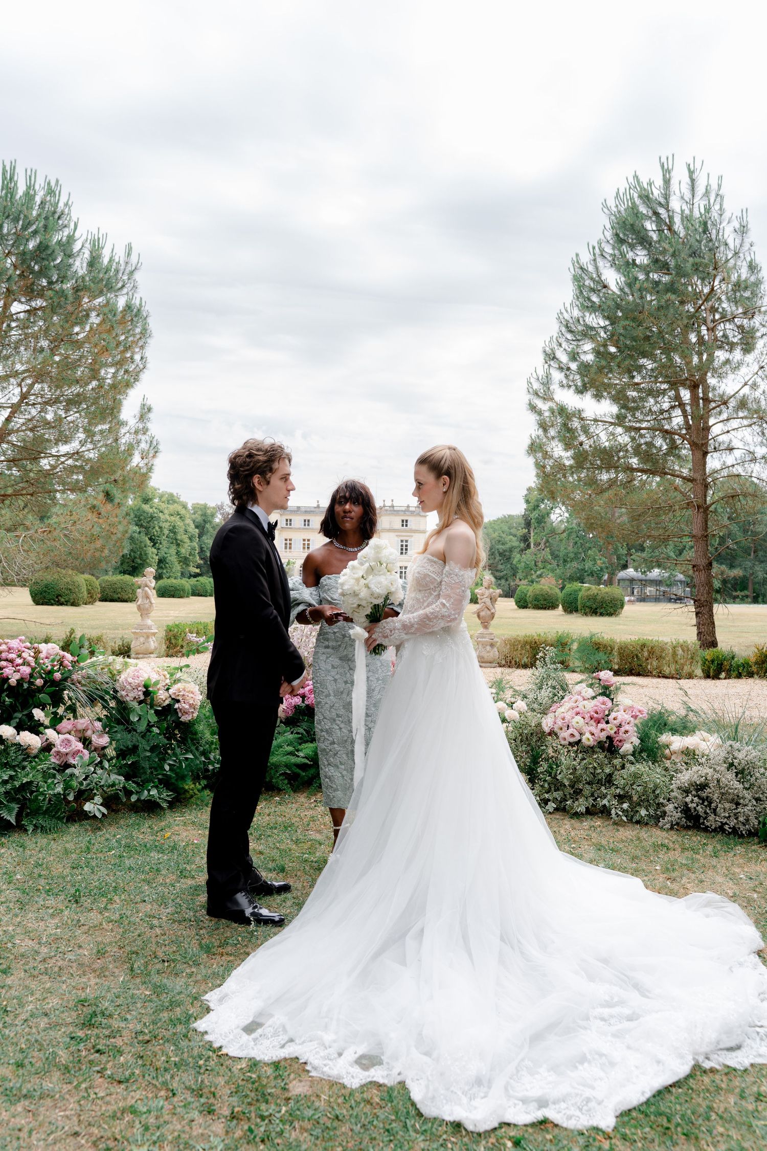 Elegant outdoor wedding ceremony with bride in lace gown and groom in tuxedo before a grand estate garden.