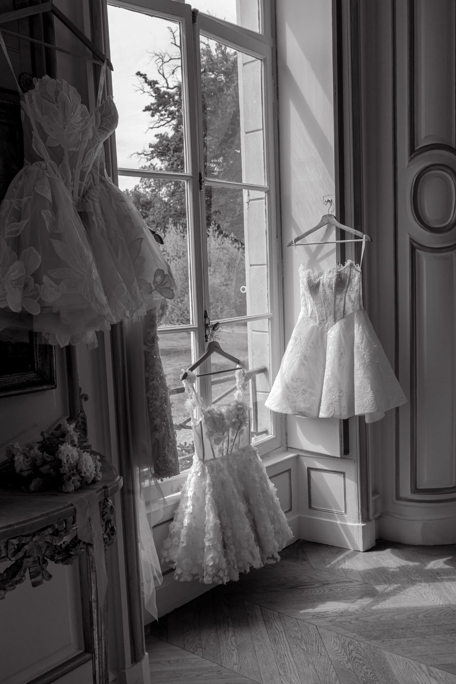 Black and white photo of elegant bridal gowns hanging by a grand window in a classic French chateau room.
