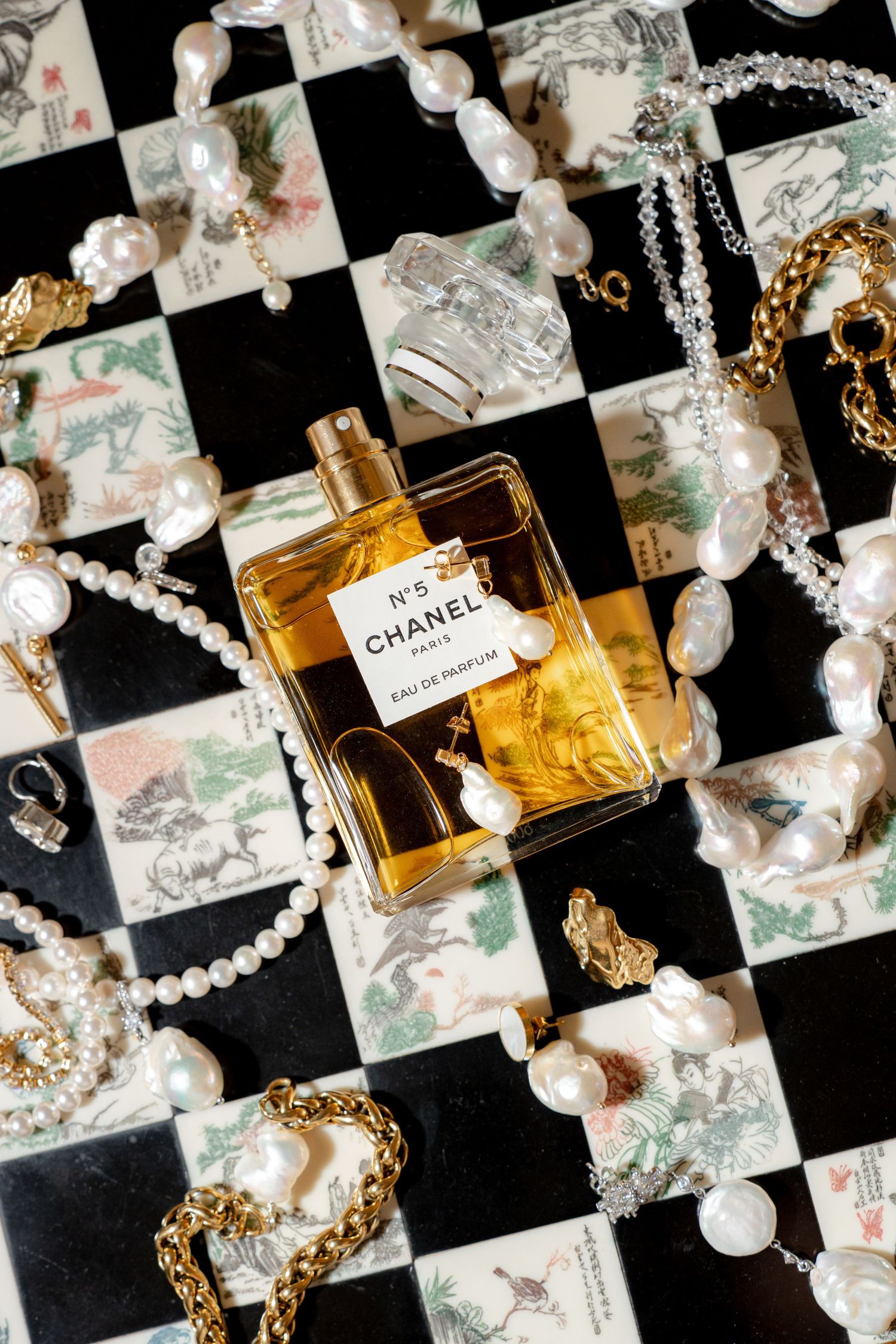 Chanel No.5 perfume bottle surrounded by pearl and gold jewelry on decorative tile surface.