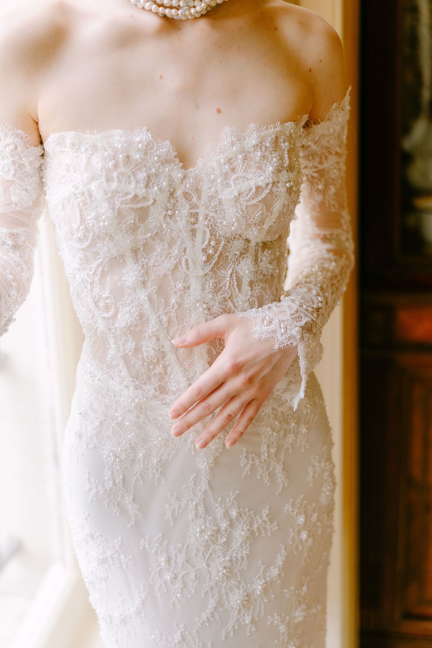 Bride in elegant off-shoulder lace wedding gown with long sleeves and pearl necklace, posing gracefully.
