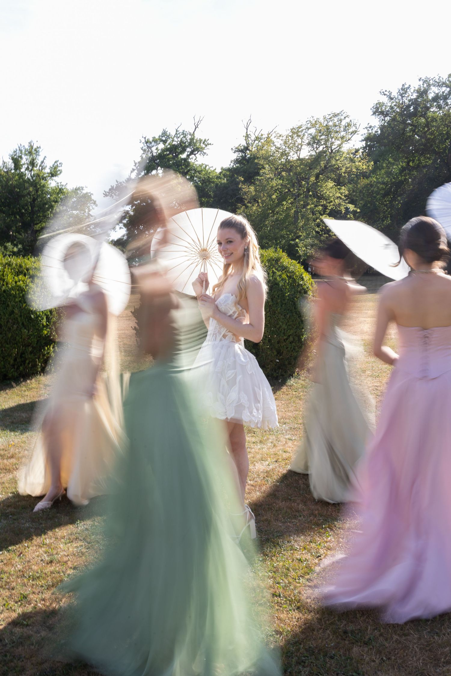 Bride in white dress holding parasol surrounded by bridesmaids in colorful gowns dancing outdoors in garden setting.
