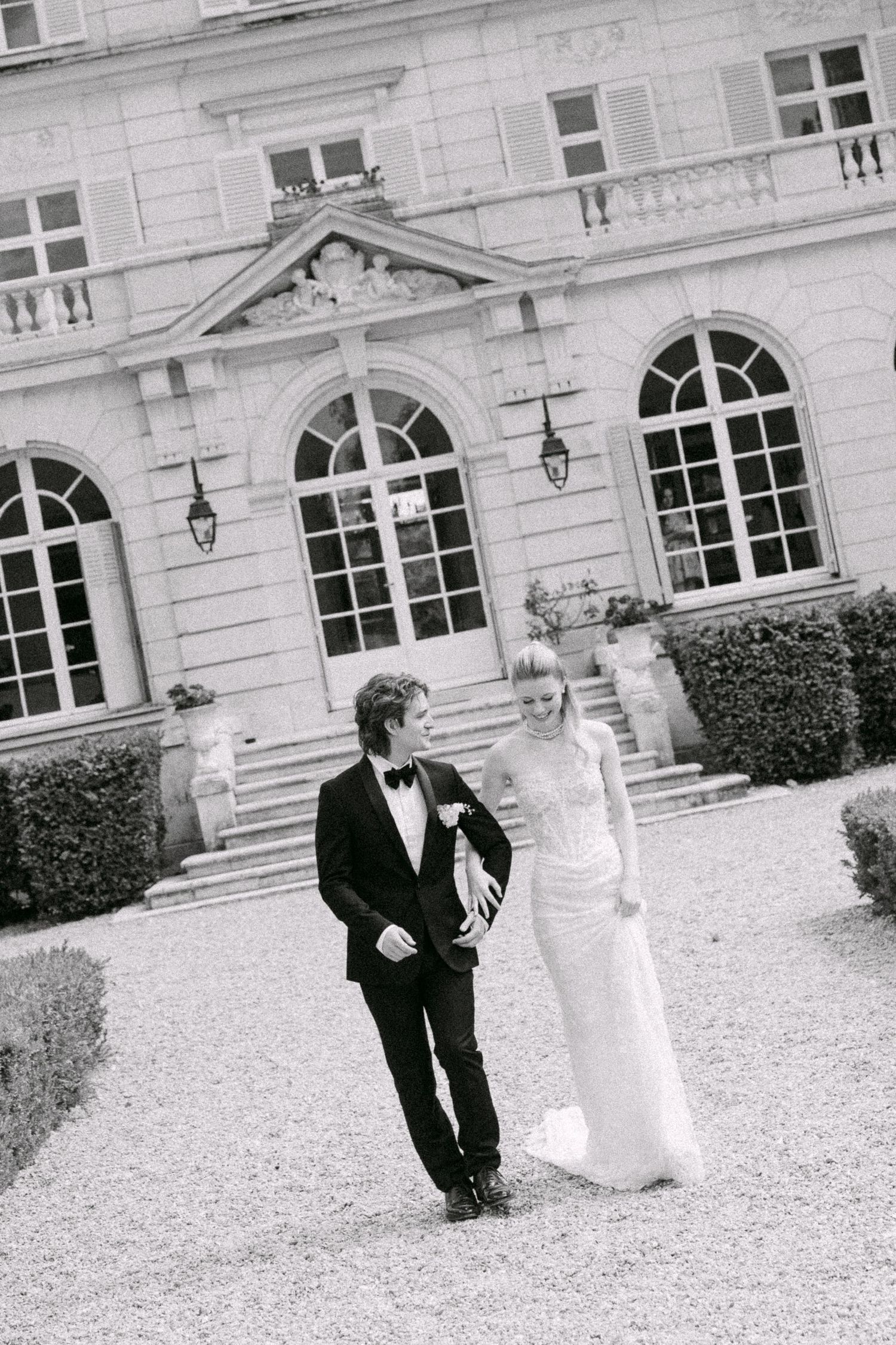 Black and white photo of a bride and groom walking outside a grand French chateau on their wedding day.