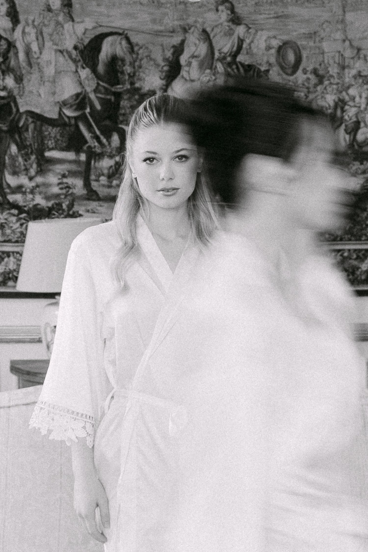 Black and white photo of a bride in white robe with blurred figure, ornate tapestry background.