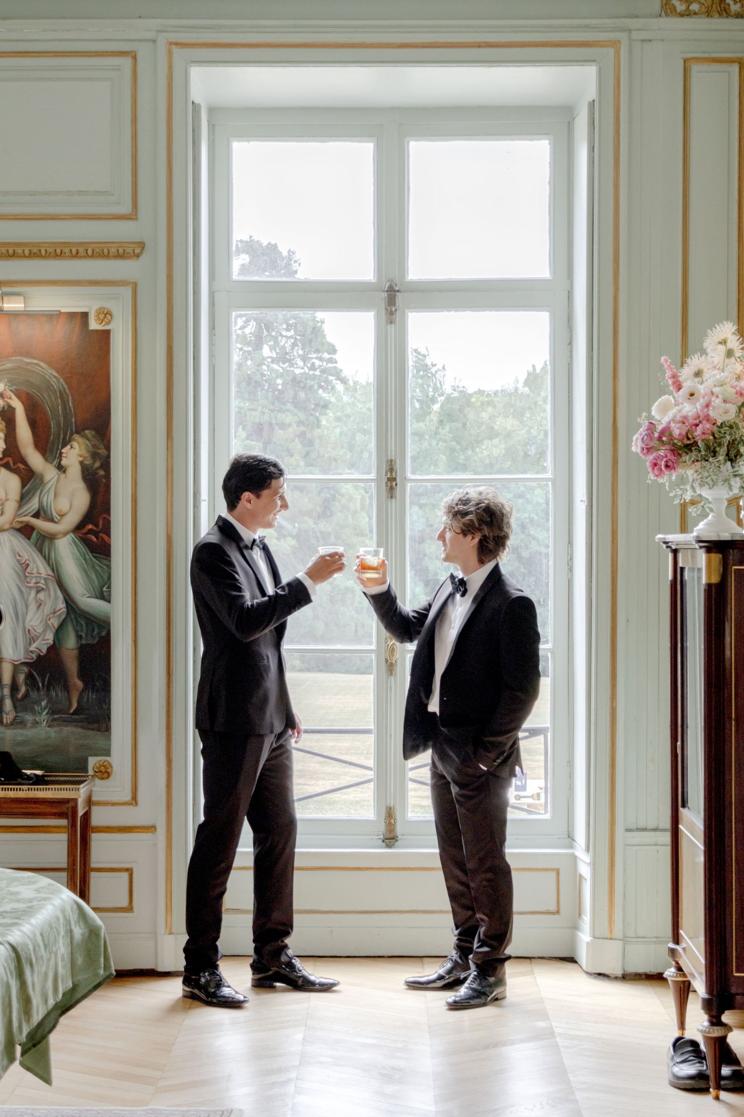 Two grooms in black tuxedos share a toast by a grand window in an elegant French-style room.