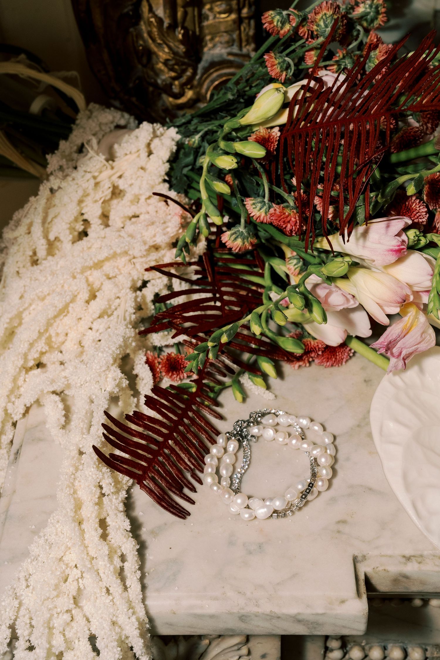 Bridal flat lay featuring pearl bracelets, dark red fern accents, tropical flowers, and white textured fabric details.