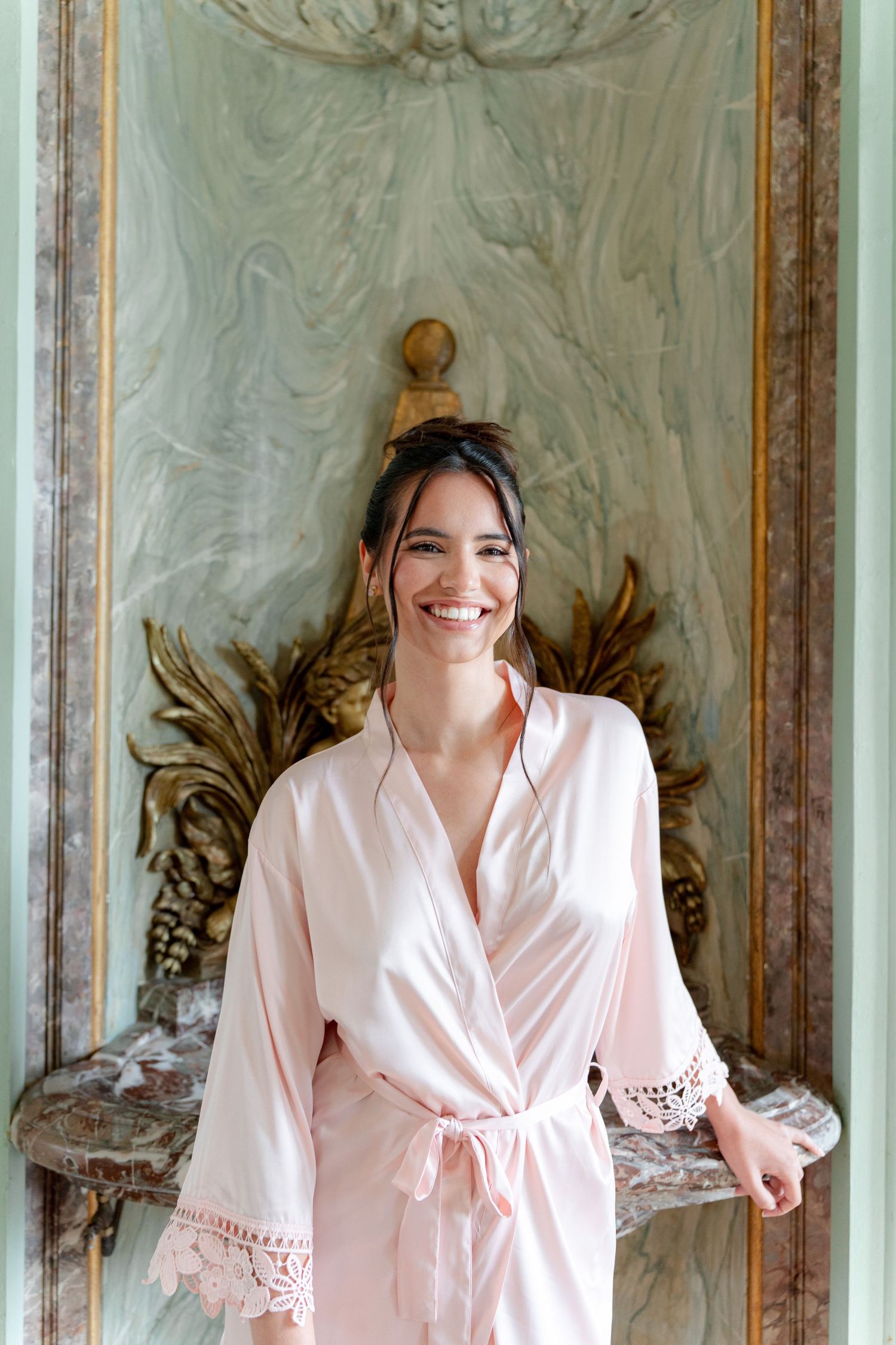 Smiling woman in pink satin lace robe standing by ornate vintage wall decor.