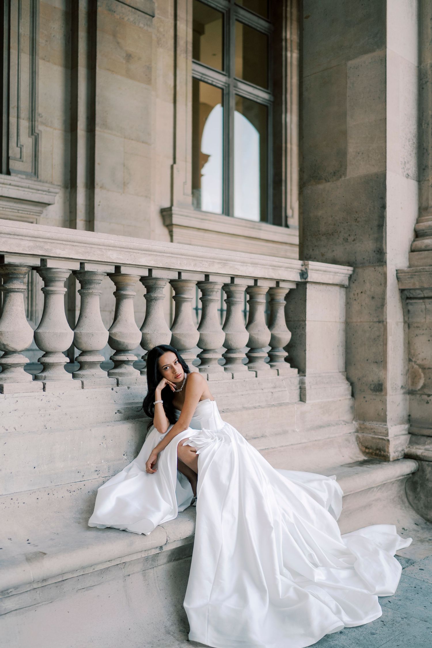 Bride in a flowing white satin gown seated on stone steps of a classical building with ornate balustrade.