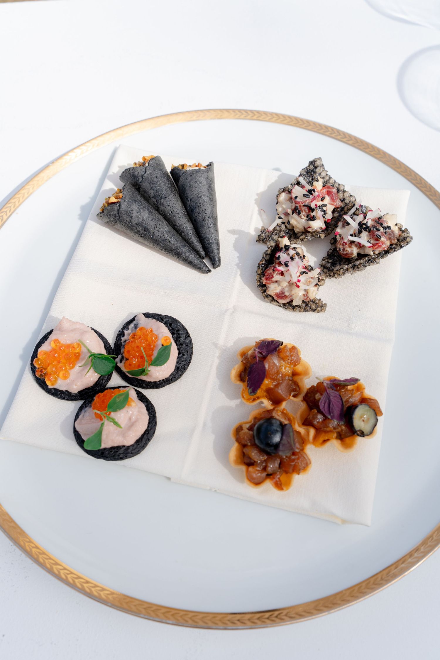 Elegant sushi platter with black seaweed cones, salmon roe rounds, sesame triangles, and garnished tartlets on gold-rimmed plate.