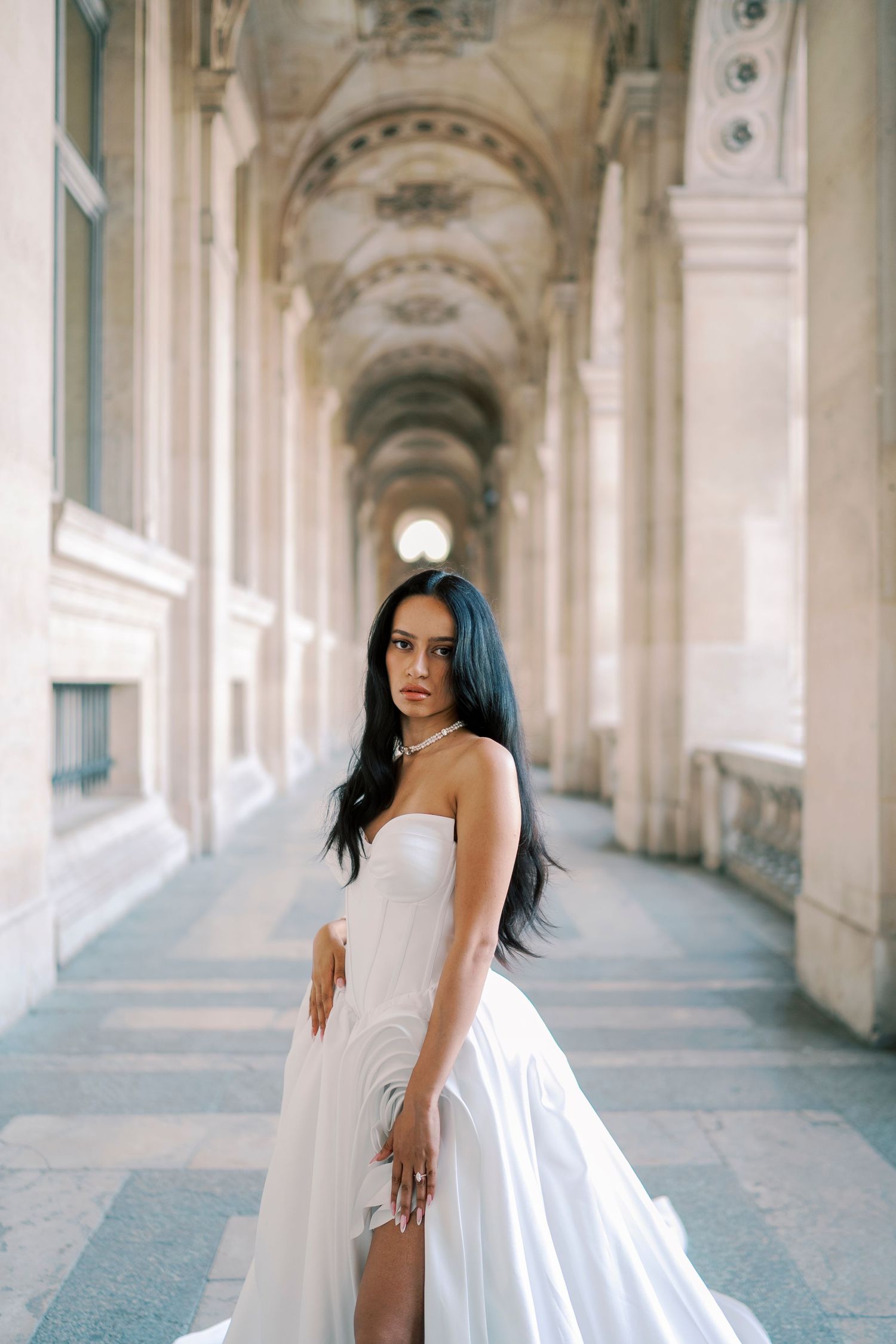Elegant bride in white strapless gown with slit poses in ornate Parisian arcade colonnade.