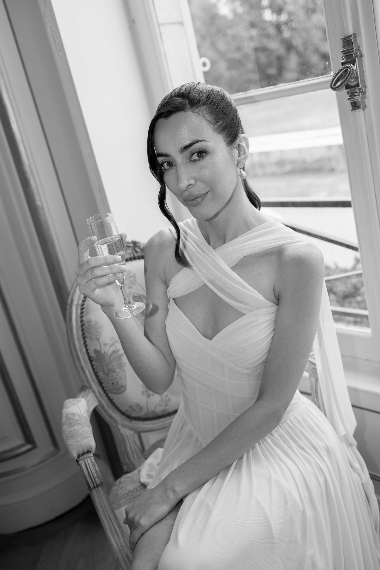 Elegant bride in white halter gown holding champagne glass, seated by window in black and white portrait photo.