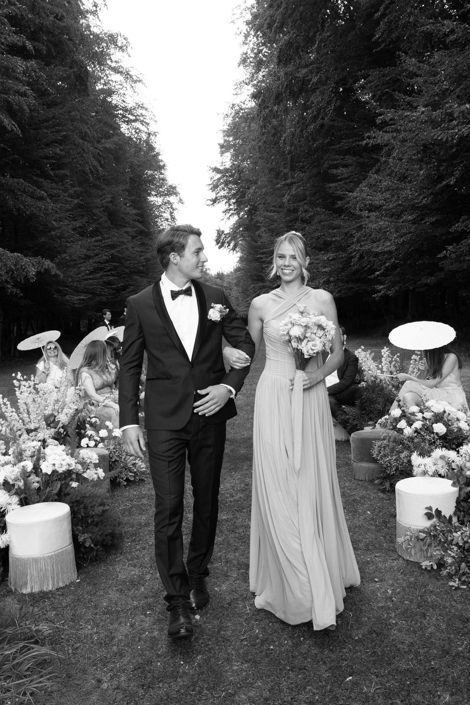 Black and white photo of a bride and groom walking down an outdoor aisle lined with flowers and guests at a wedding.