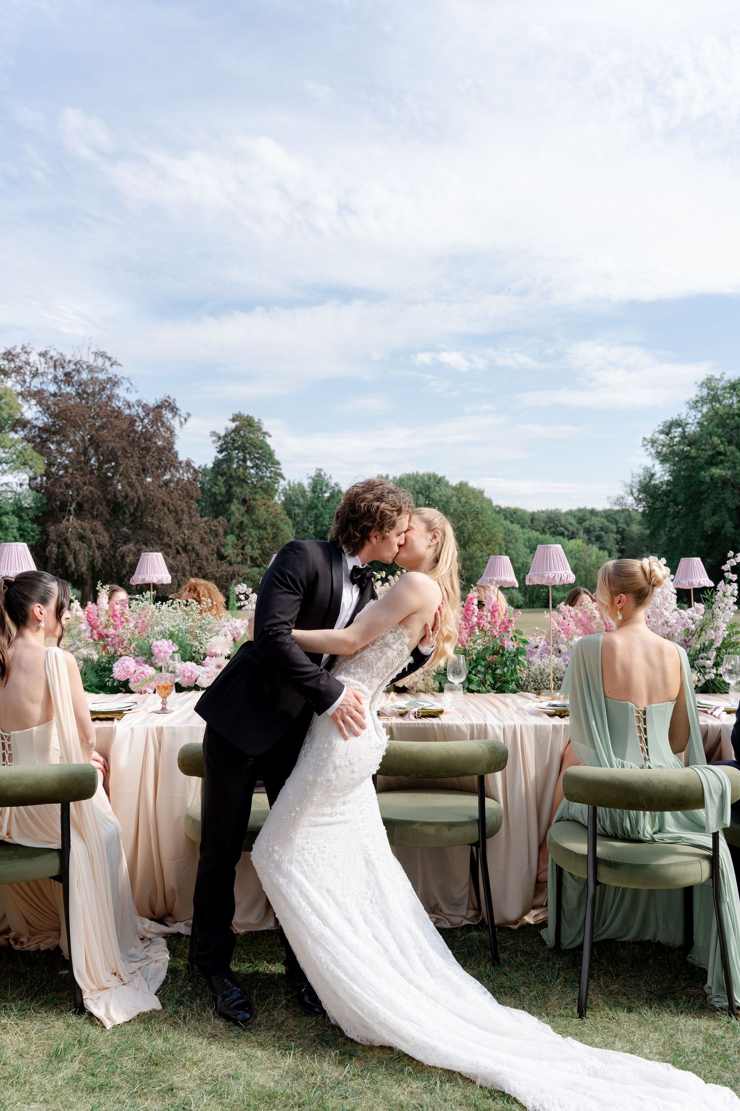 Bride and groom share a kiss at an elegant outdoor wedding reception with pink floral arrangements and green velvet chairs.