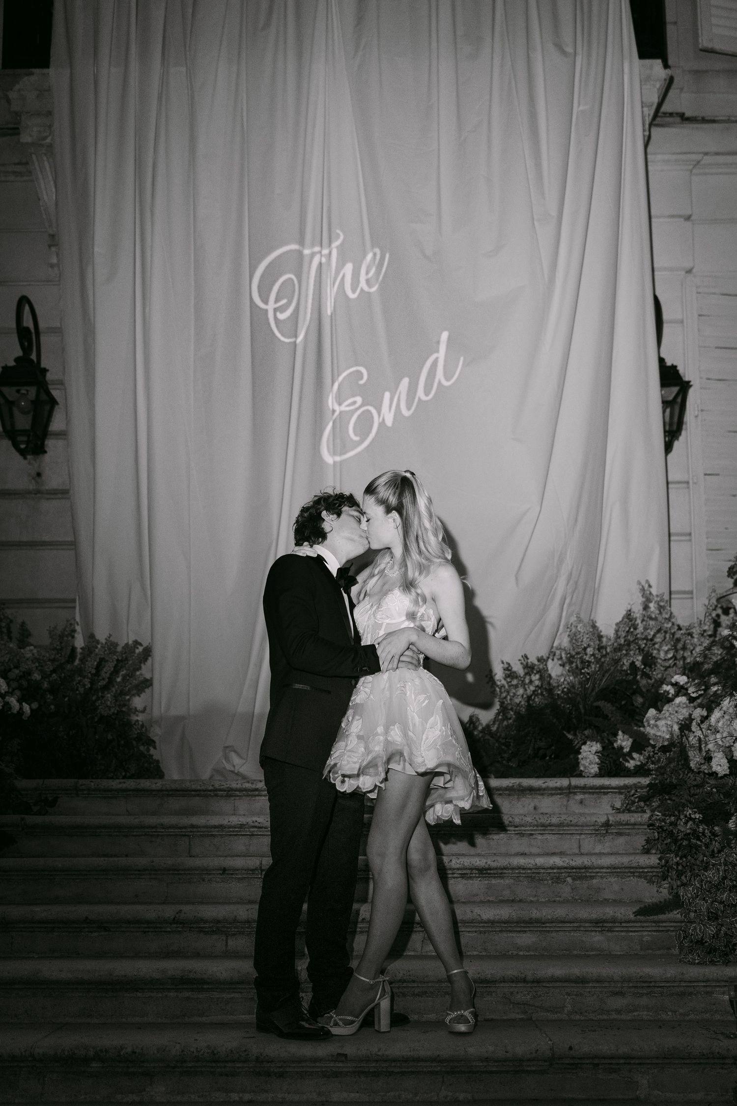 Black and white photo of a couple kissing on steps before a draped backdrop with neon sign reading The End.