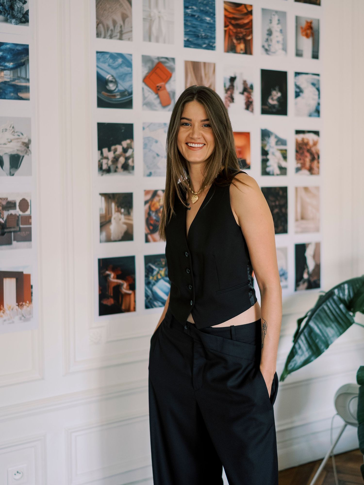 Smiling woman in black sleeveless top and wide-leg pants poses in front of a colorful photo mood board wall.