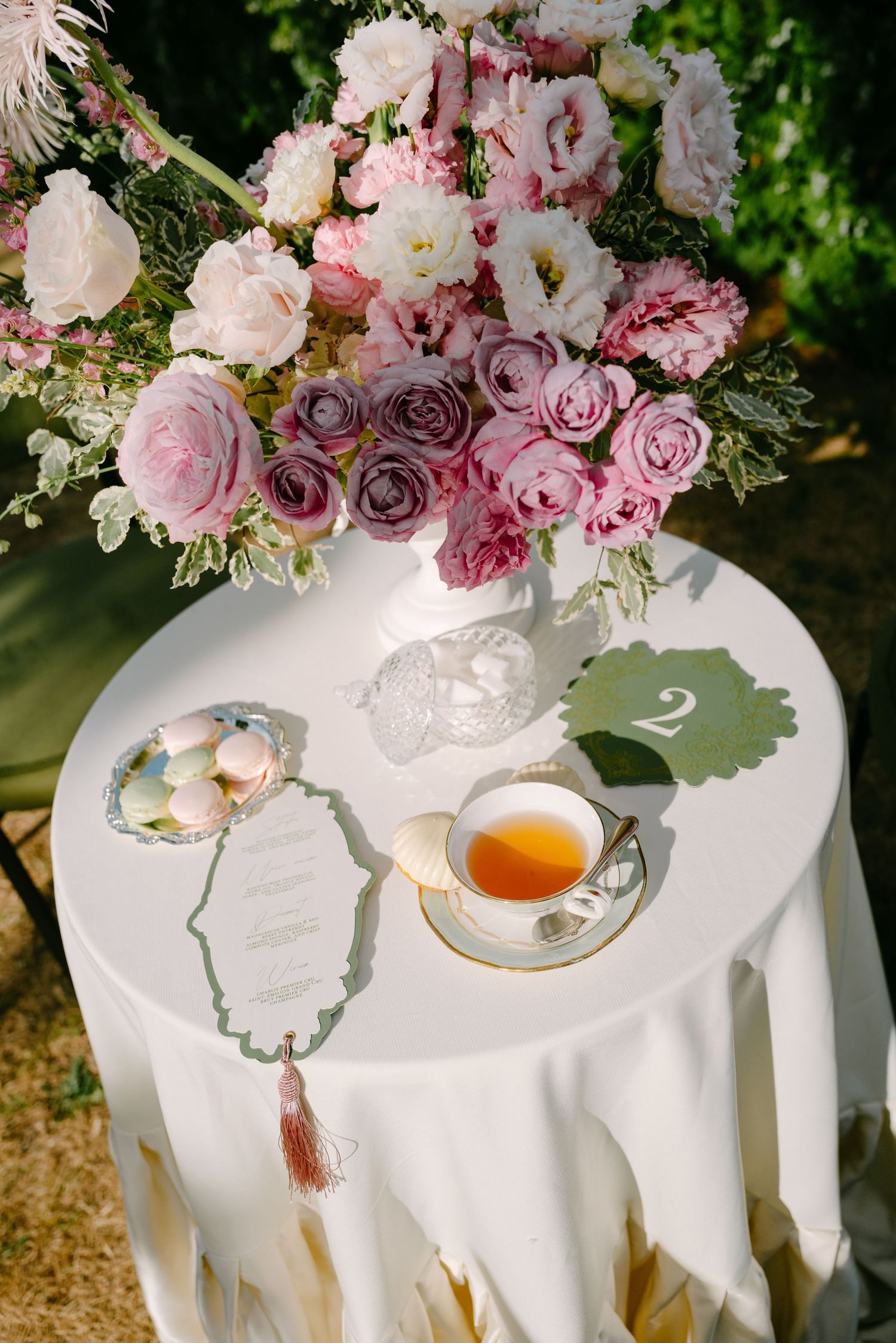 Elegant wedding table with pink floral arrangement, tea cup, macarons, and menu card on white tablecloth.