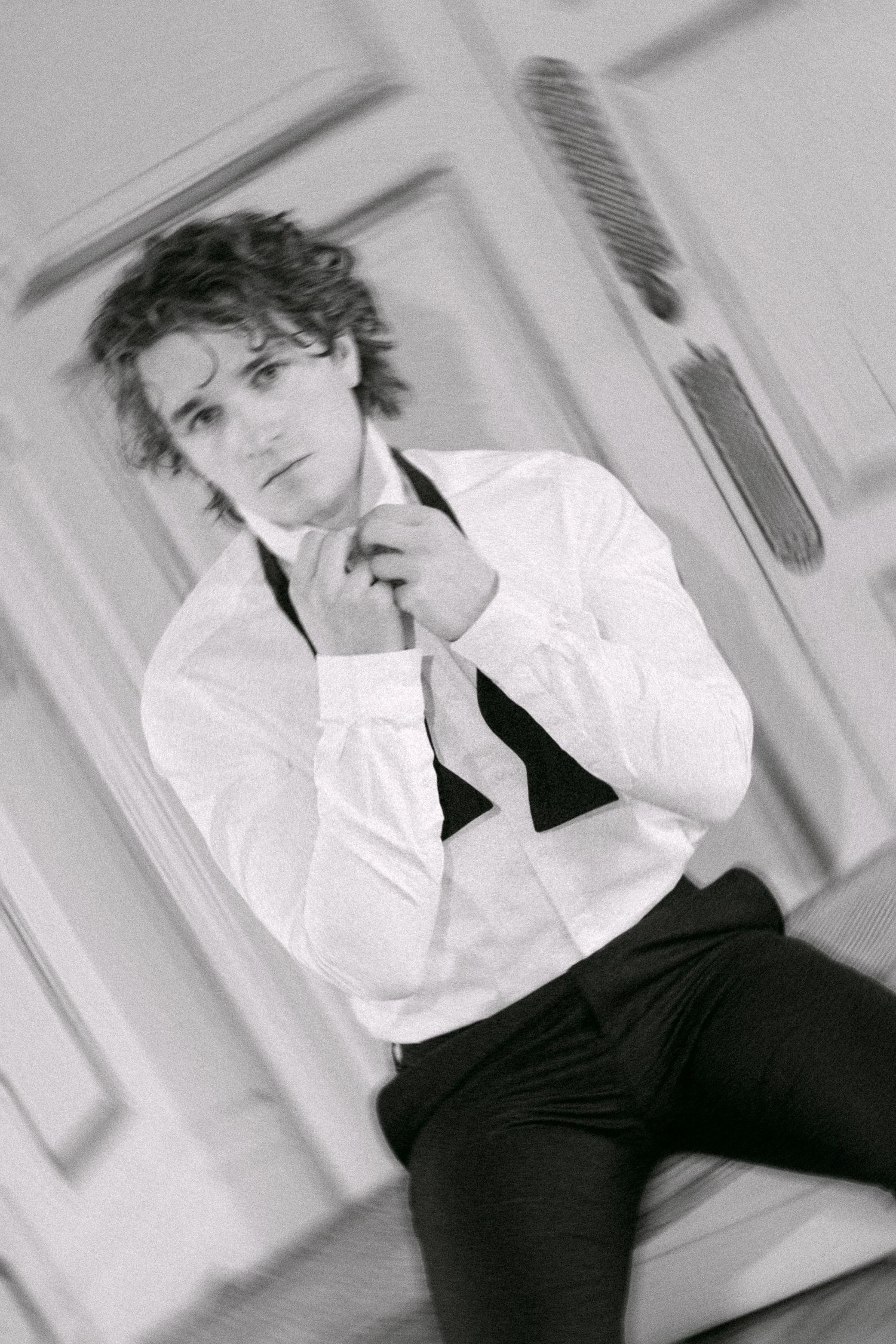 Black and white photo of a young man in white dress shirt adjusting his bow tie, sitting in an elegant room.