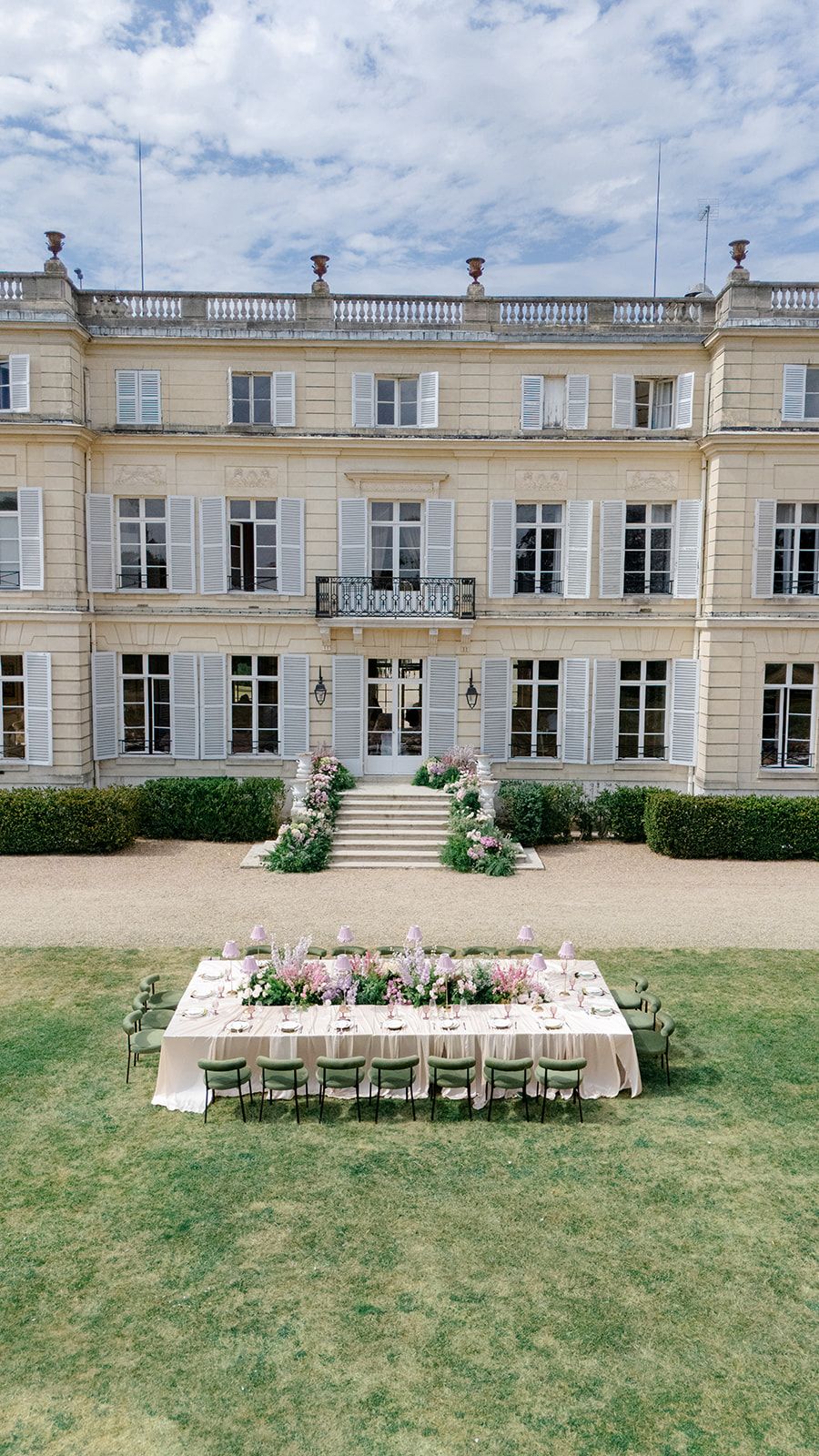 Outdoor wedding reception table set before grand French chateau with floral centerpieces.