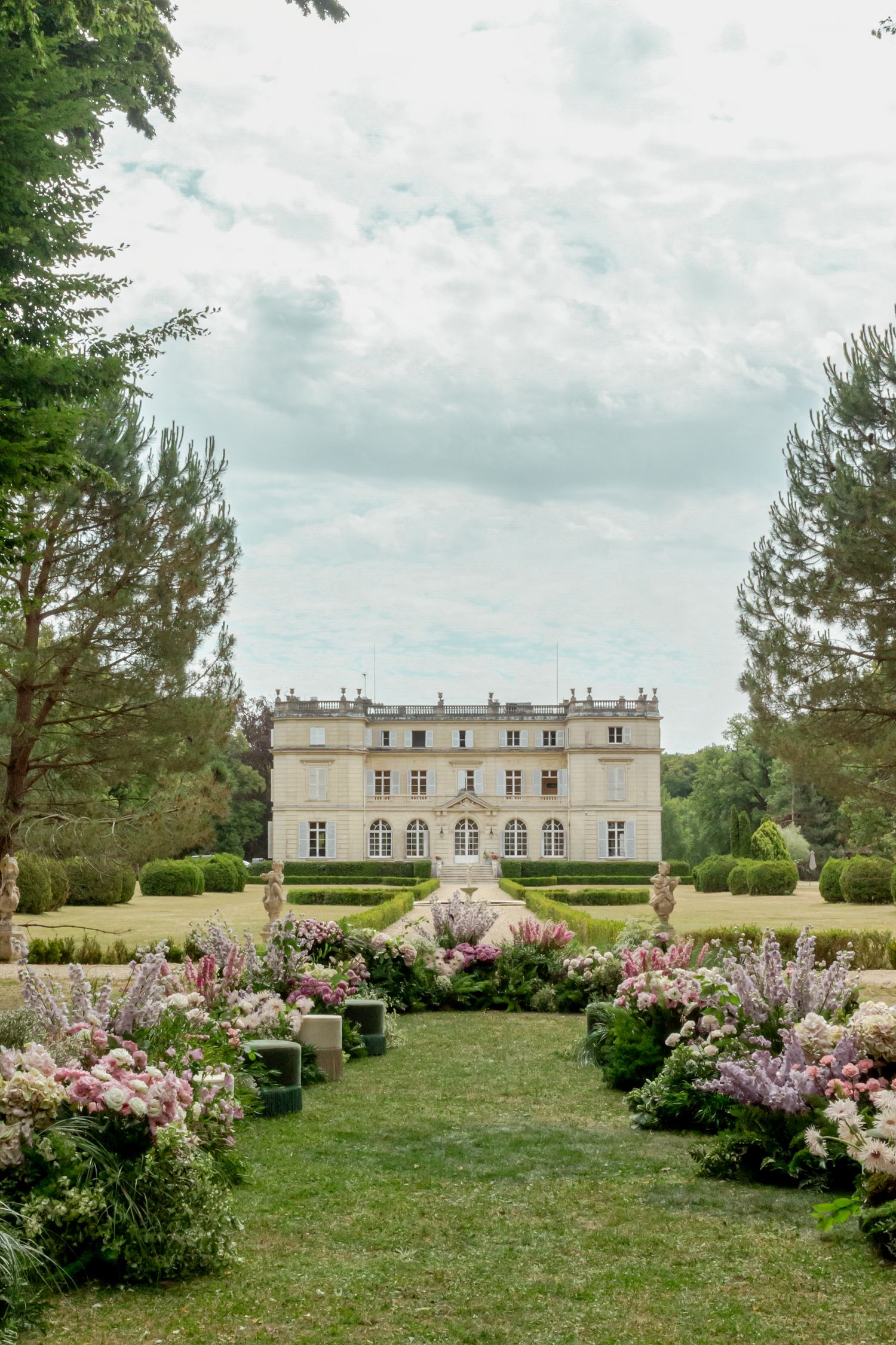 Elegant French château wedding venue with lush floral arrangements lining a garden aisle under a cloudy sky.