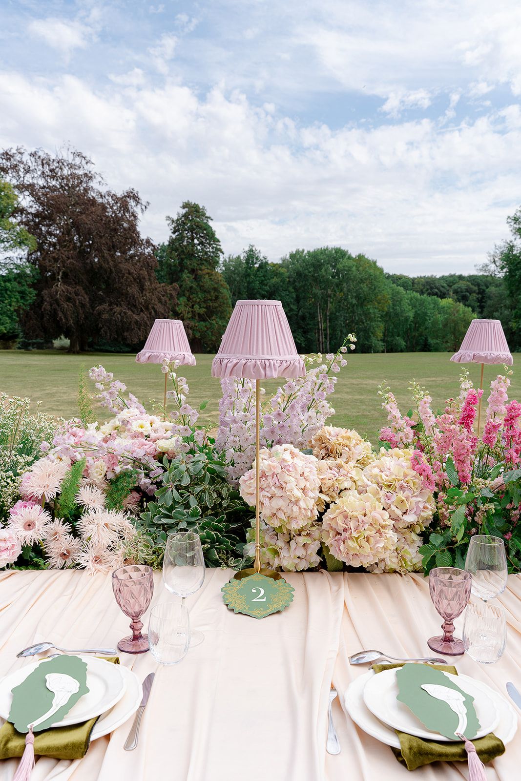 Elegant outdoor wedding table with pink lamps, hydrangeas, and greenery under a cloudy sky.