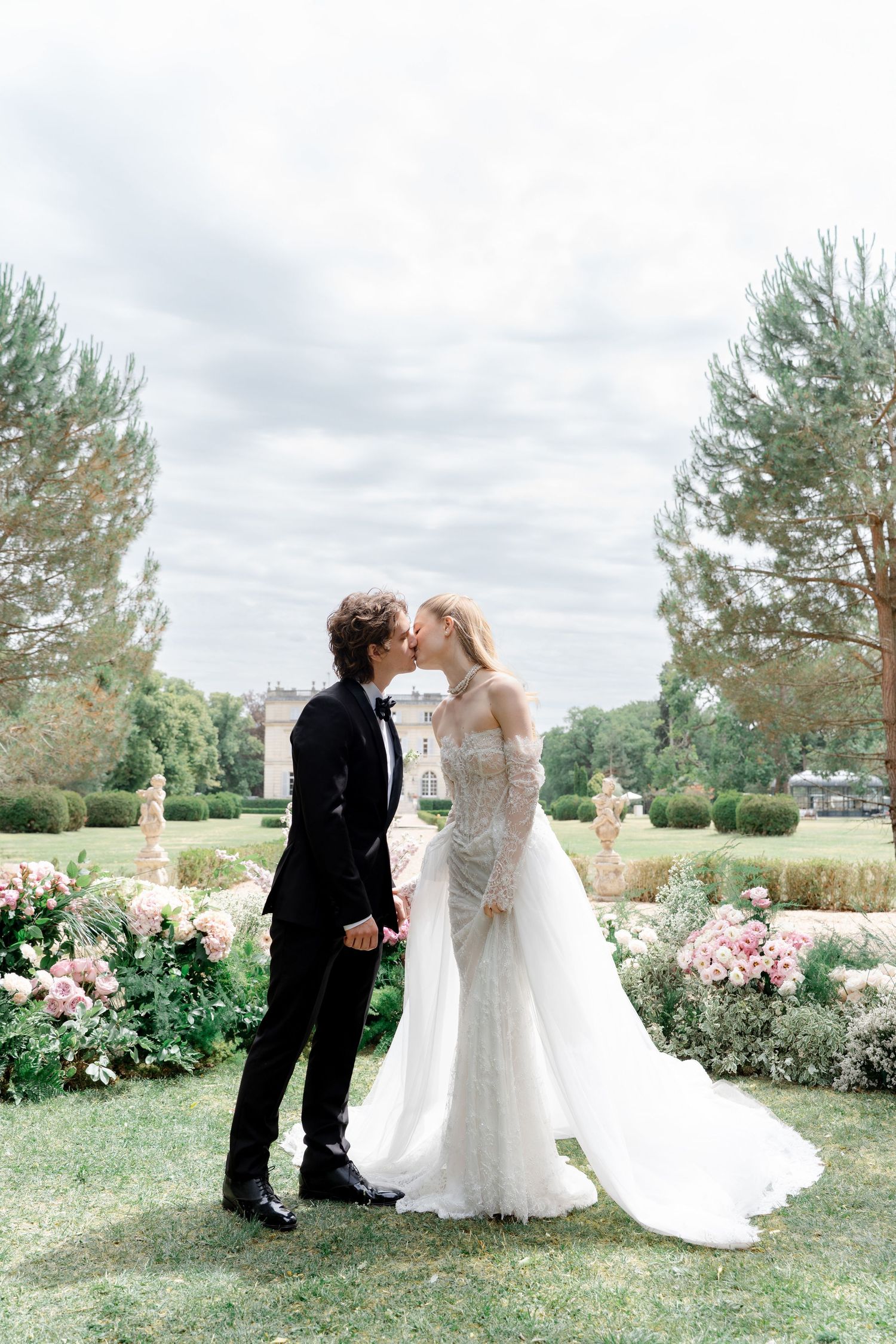 Bride and groom sharing a kiss in elegant wedding attire at a French château garden with roses and manicured hedges.