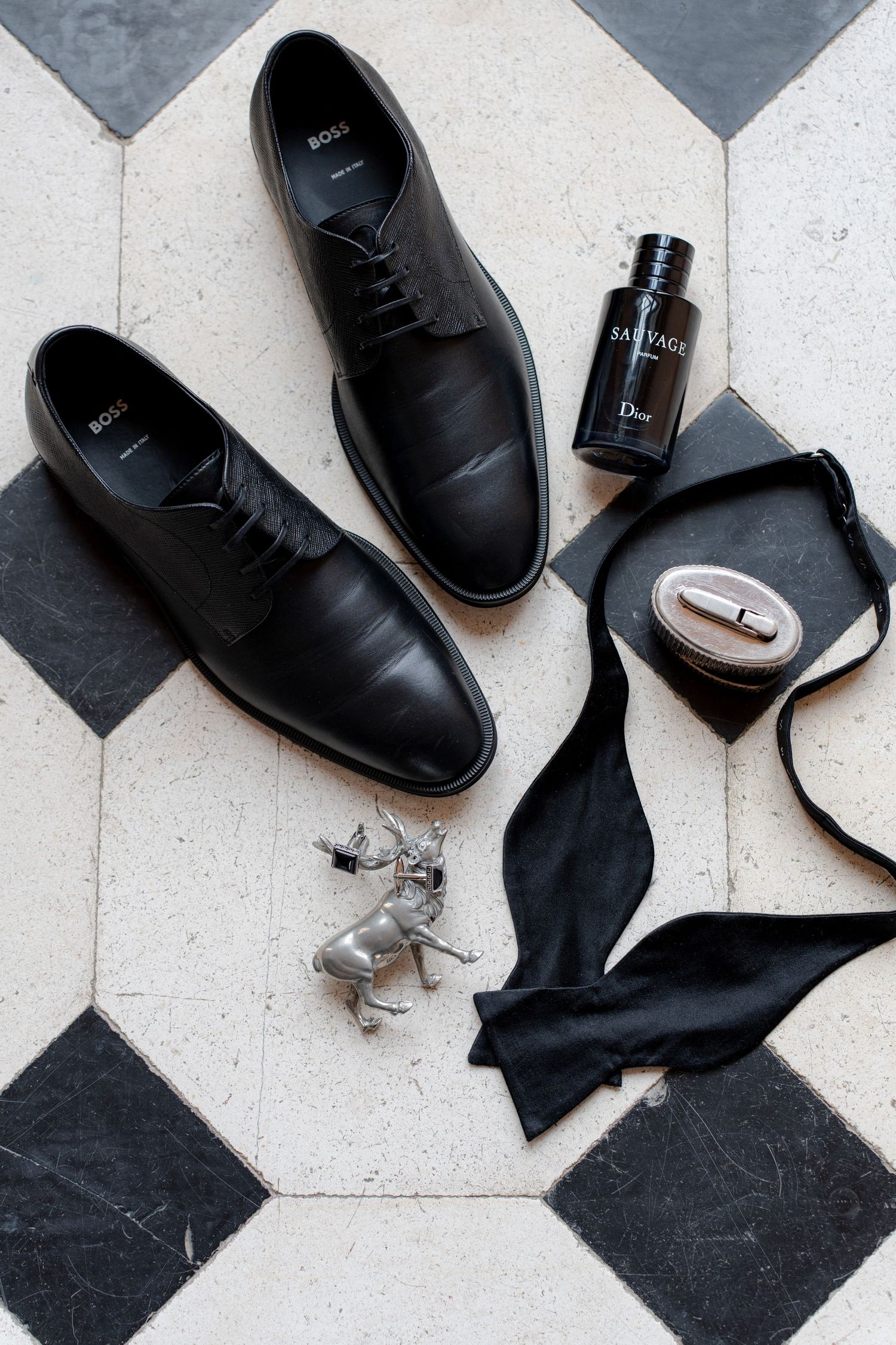 Black Hugo Boss dress shoes with Dior Sauvage cologne, bow tie, cufflinks, and silver deer figurine on geometric tile floor.