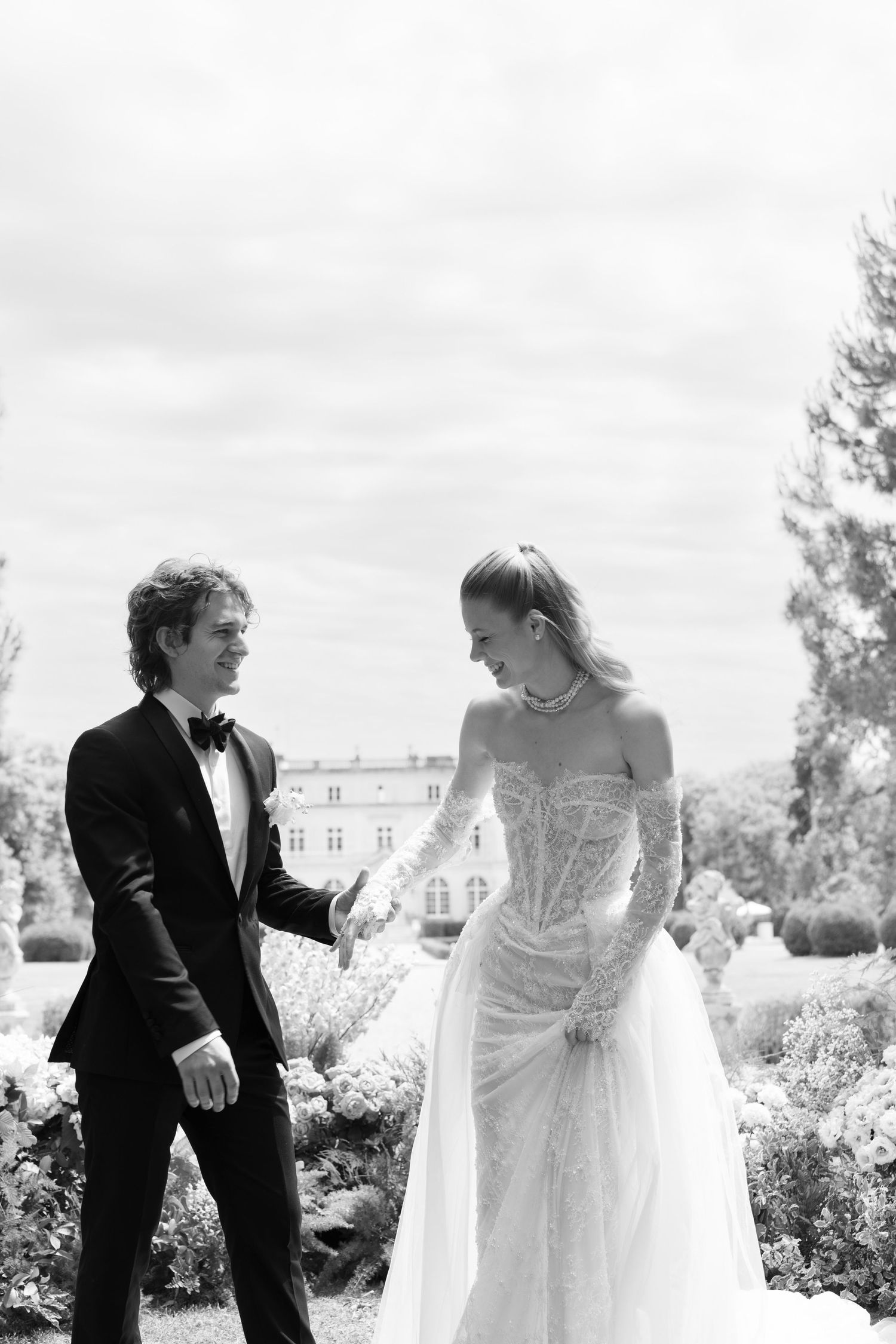 Black and white photo of a bride in a lace gown and groom in a tuxedo holding hands in a garden.