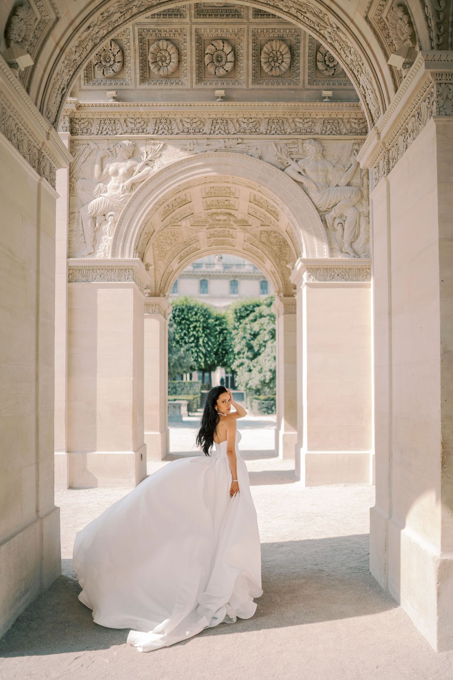 Bride in flowing white gown poses under ornate stone archway of classical European building.