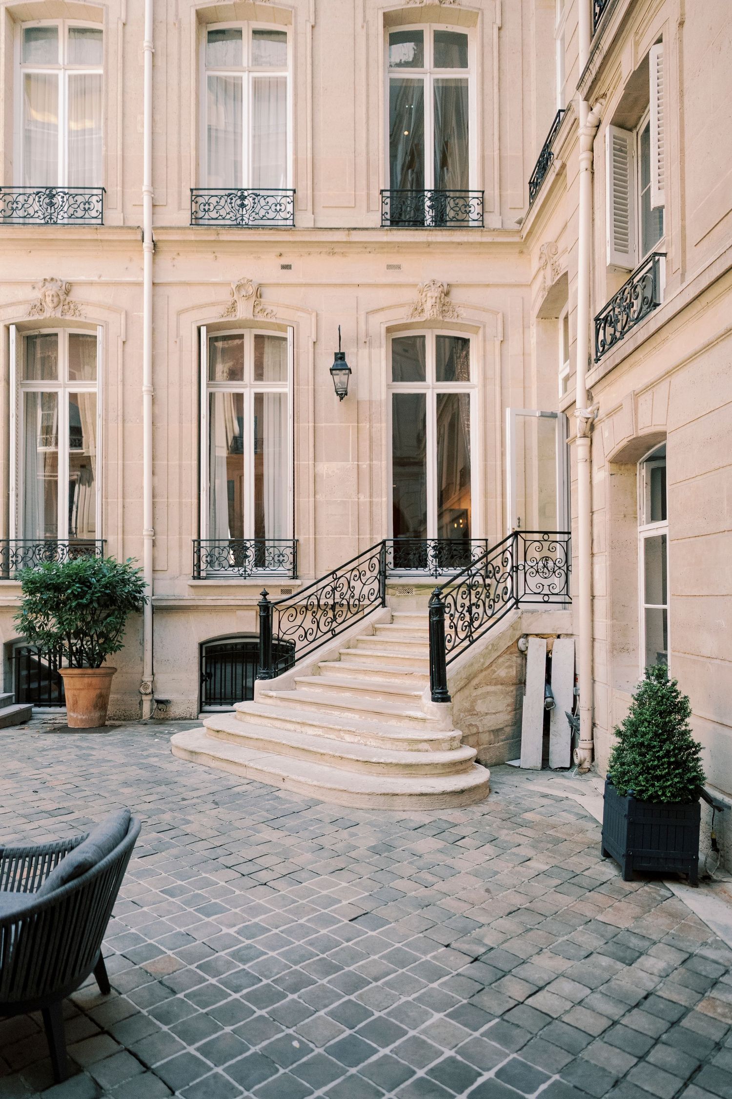 Elegant Parisian courtyard with ornate wrought iron staircase, tall windows, and potted topiaries on cobblestone pavement.