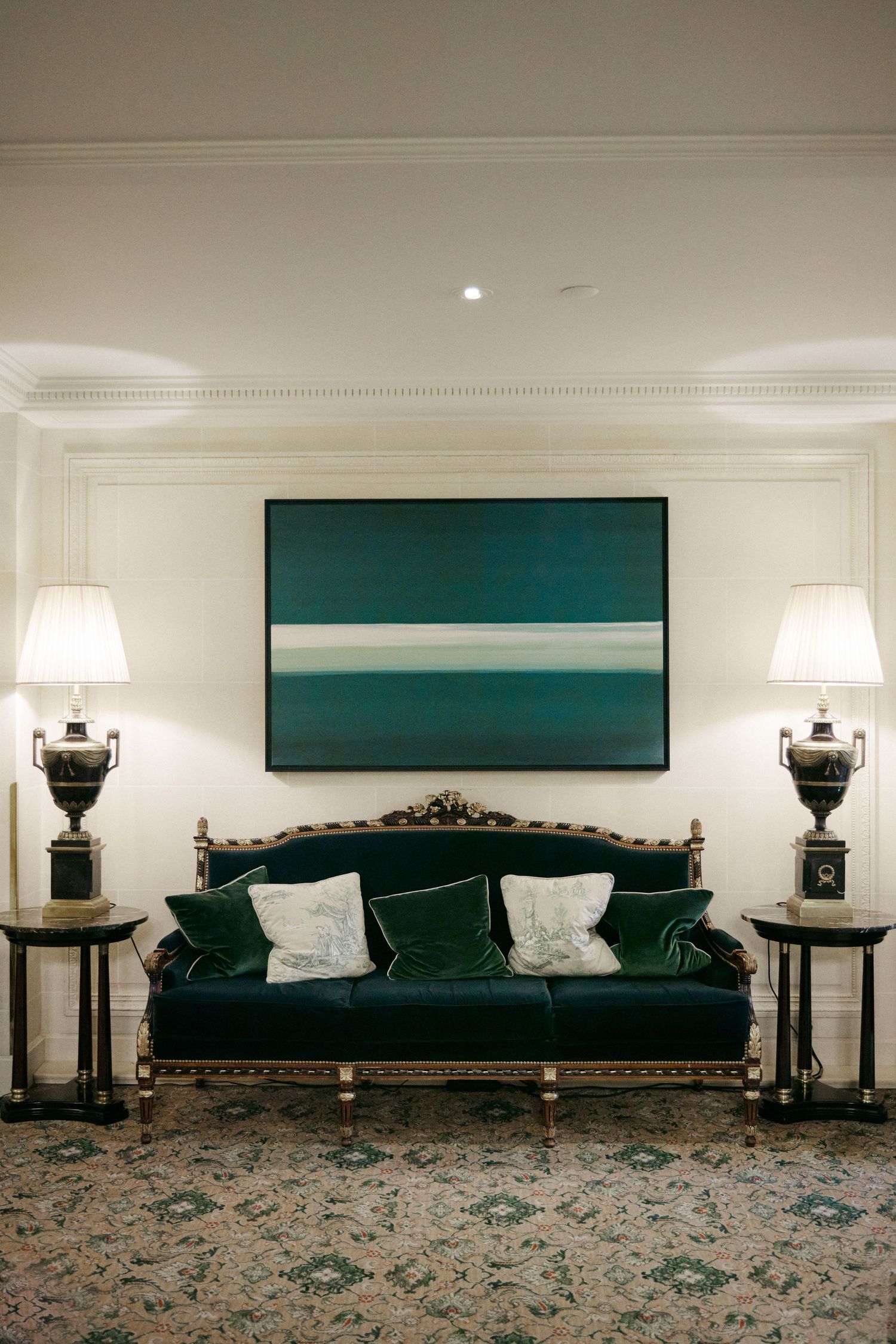 Elegant green velvet sofa with gold frame, flanked by lamps, beneath a teal abstract painting in a classic interior.